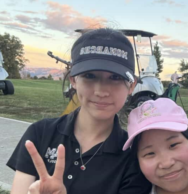 Two young girls outdoors at sunset, one wearing a black visor with 'MORATOR' written on it and making a peace sign, the other wearing a pink cap and smiling.