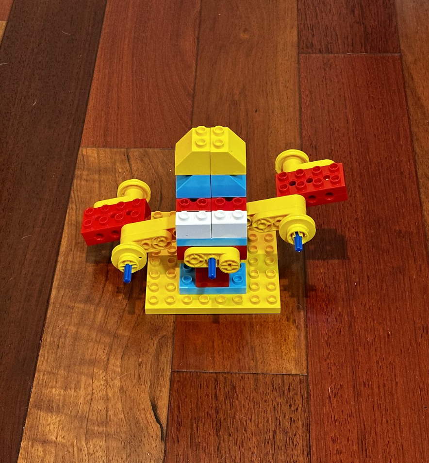 LEGO construction of a small colorful spacecraft with yellow, red, blue, and white bricks, placed on a wooden floor.