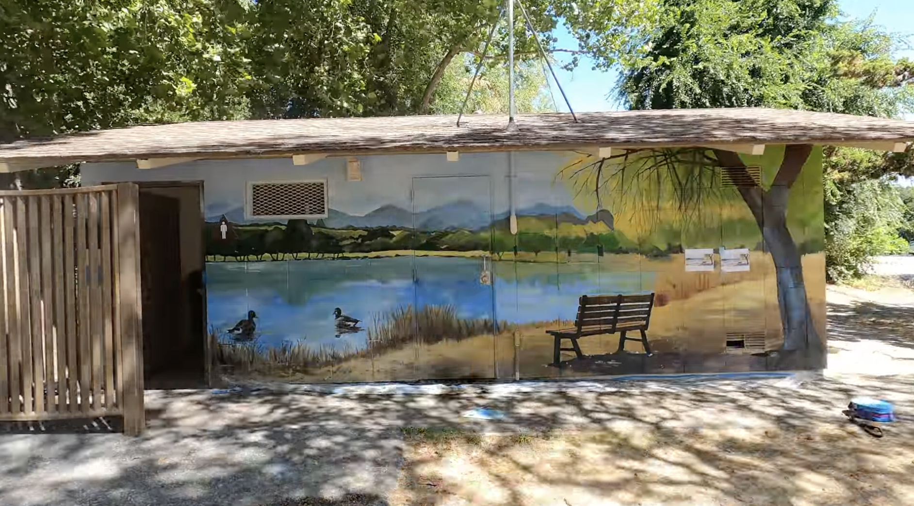 Building with landscape mural depicting a lake, bench, ducks, and trees; surrounded by real trees and outdoor setting.