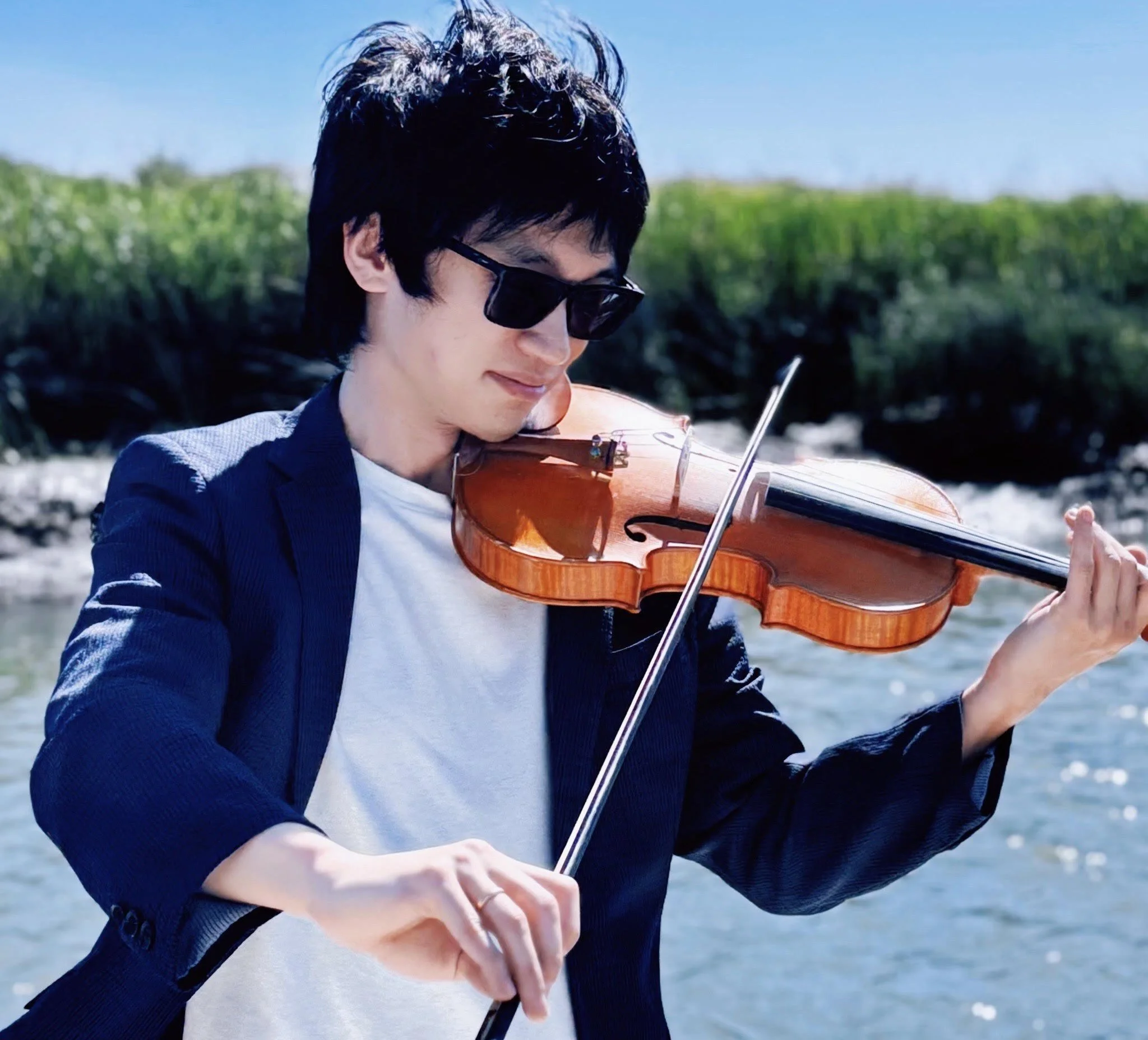 A young man wearing sunglasses and a blue blazer playing the violin outdoors near a river with green bushes in the background.