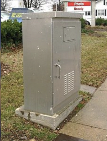Metal electrical utility box on sidewalk with grass and sign in background.
