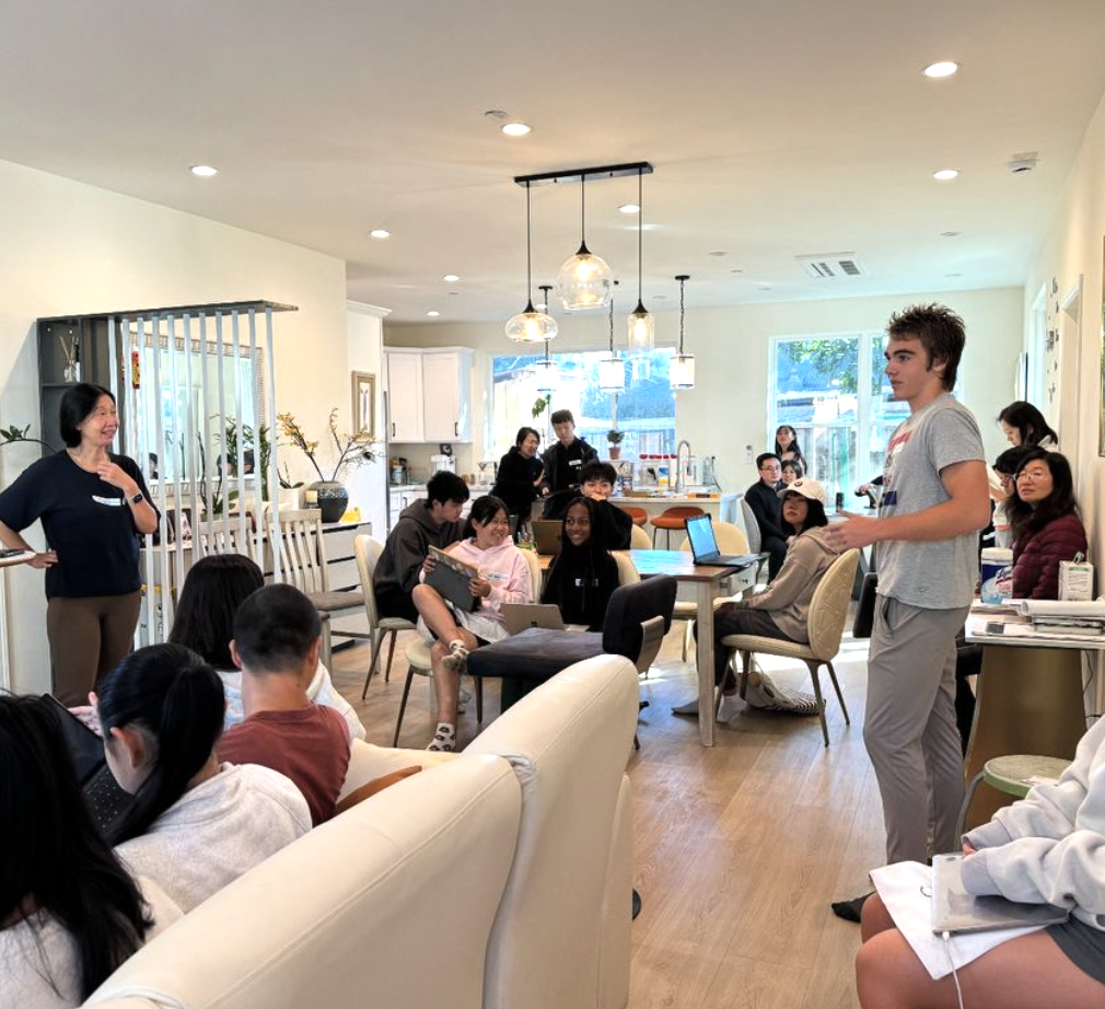 A group of people gathered in a living room for a meeting, some seated on couches and chairs, others standing. A person is speaking while others listen attentively. Laptops and notebooks are visible.