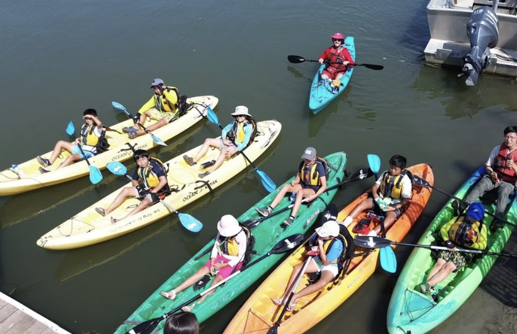 A group of people sitting in kayaks on water, wearing life jackets and holding paddles.