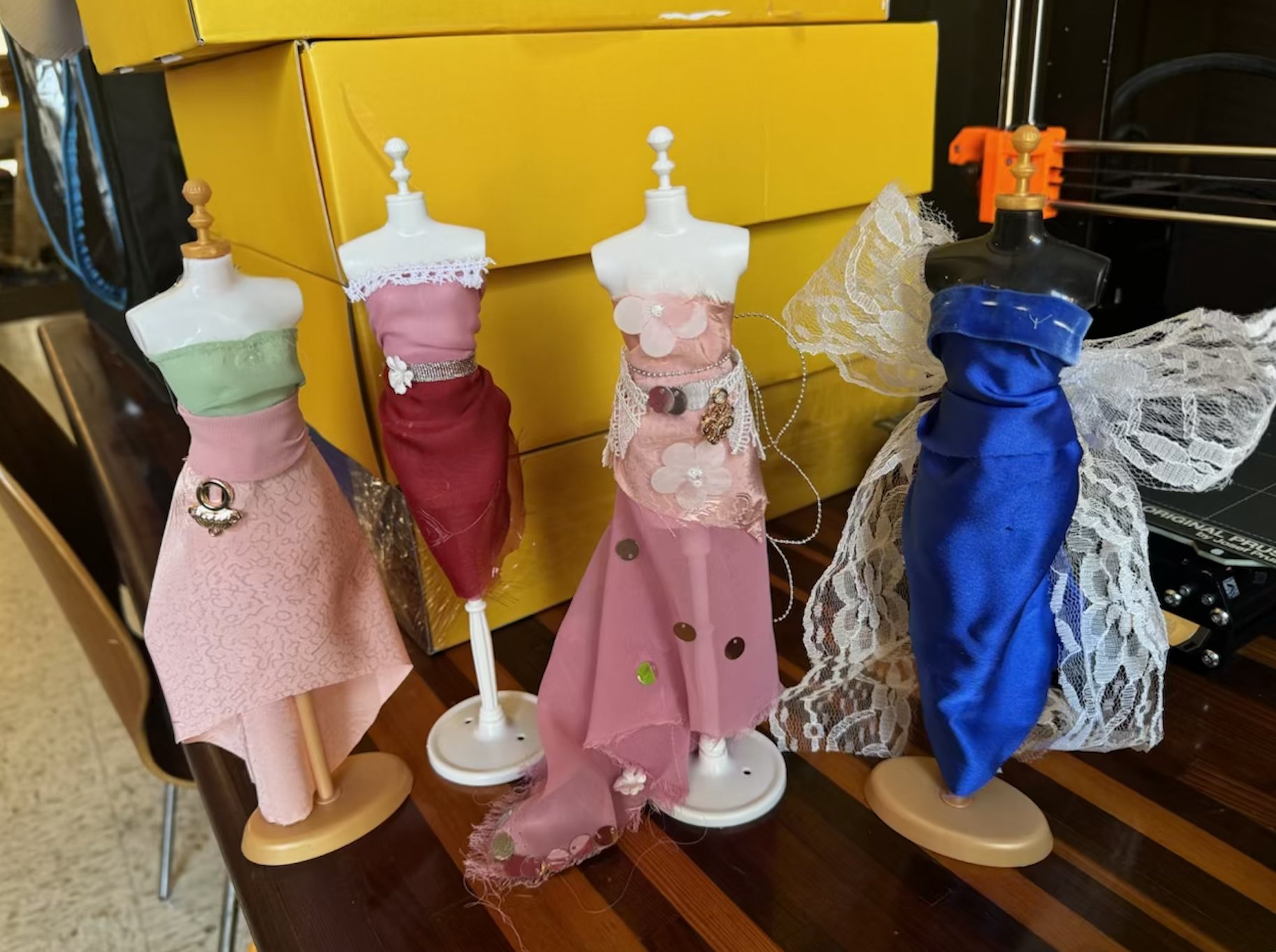 Four miniature dress mannequins displaying various colorful fabric designs, including pink, red, and blue with lace and decorative embellishments, placed on a wooden table in front of yellow boxes.