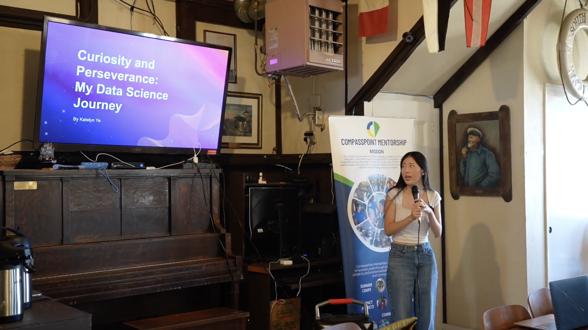 Person giving a presentation beside a screen displaying "Curiosity and Perseverance: My Data Science Journey" in a room with various equipment and a mentorship banner.