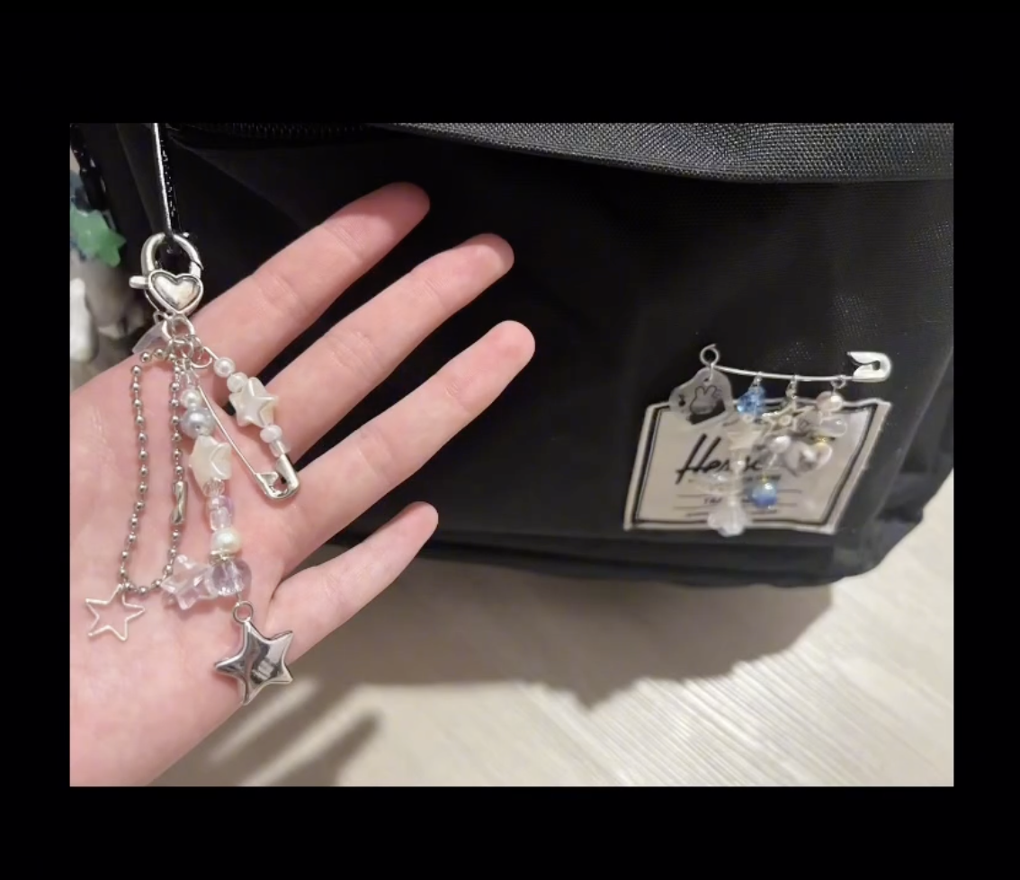 Black backpack with decorative charms, including stars and beads, attached to it. A hand holding a similar star charm is also visible.