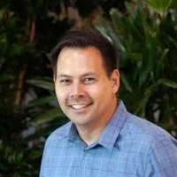 A man with short dark hair smiling outdoors, wearing a blue checkered shirt, with green foliage in the background.