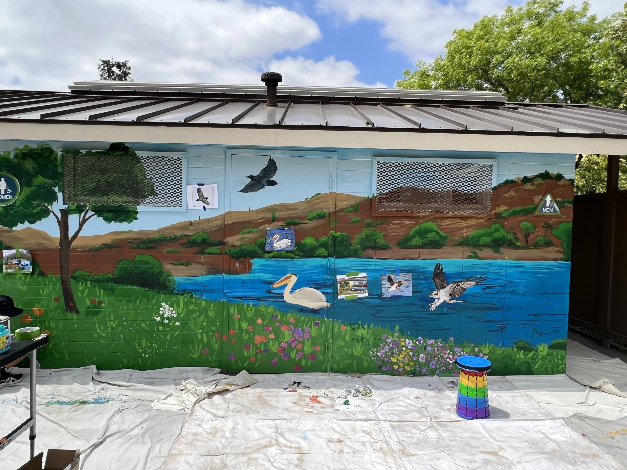 A colorful mural on a building wall depicting a natural scene with a lake, hills, and birds. Trees and flowers are painted in the foreground near the bottom, and the sky is partly cloudy. The mural is still in progress, with paint supplies and tarps on the ground. Wall signs indicate men's and women's restrooms.