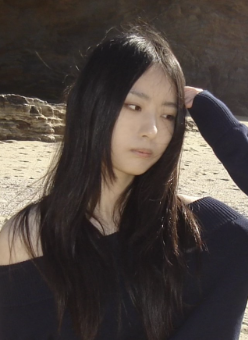 Young woman with long black hair looking down and to the side outdoors, with a rocky background.