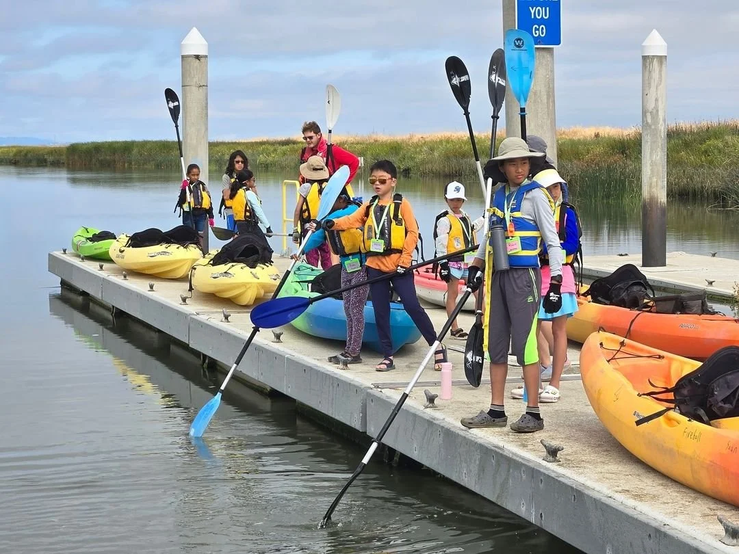 A group of people wearing life vests holding paddles, standing on a dock with kayaks in the water.