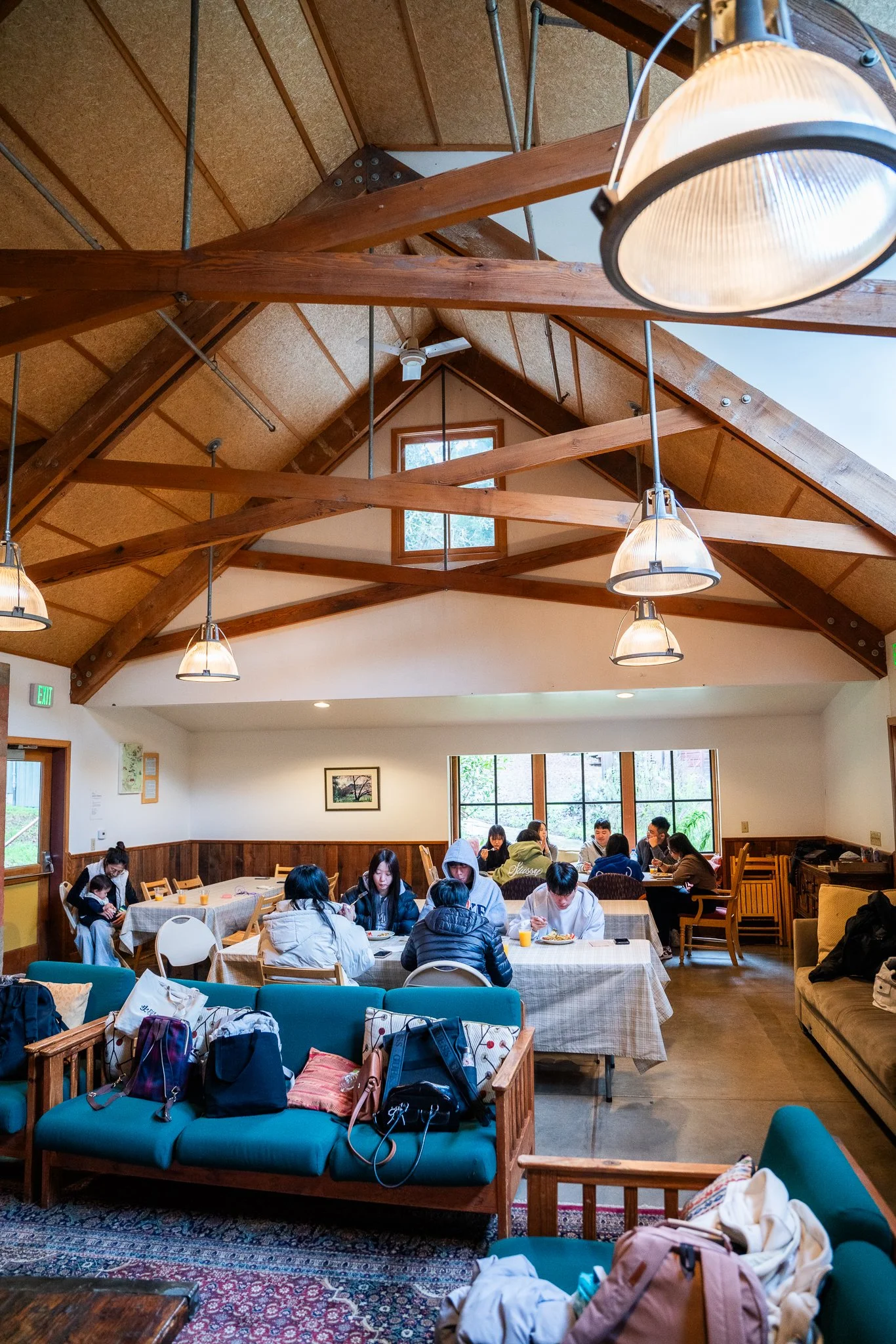 Interior of a cozy wooden dining room with people seated at tables, enjoying meals and conversing. The room features exposed wooden beams, large windows, and ambient lighting. Bags and coats are placed on couches in the foreground.