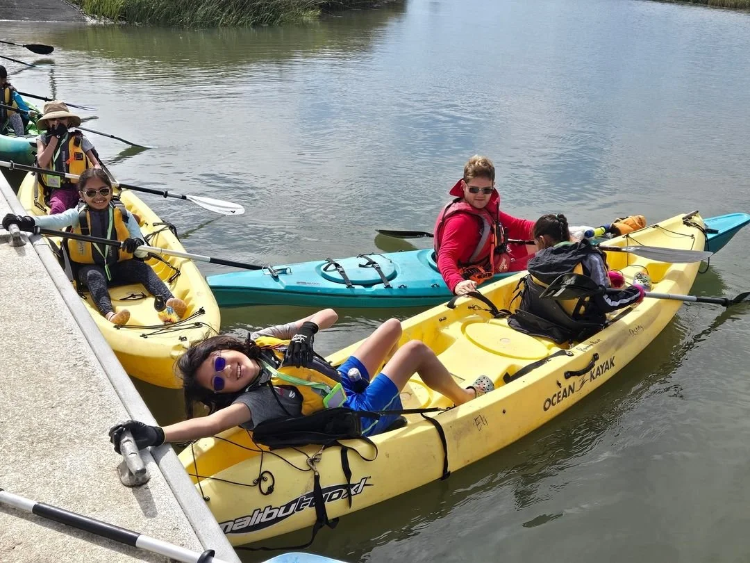 Group of people enjoying a kayaking trip on a lake, wearing life jackets, and posing in yellow and blue kayaks.