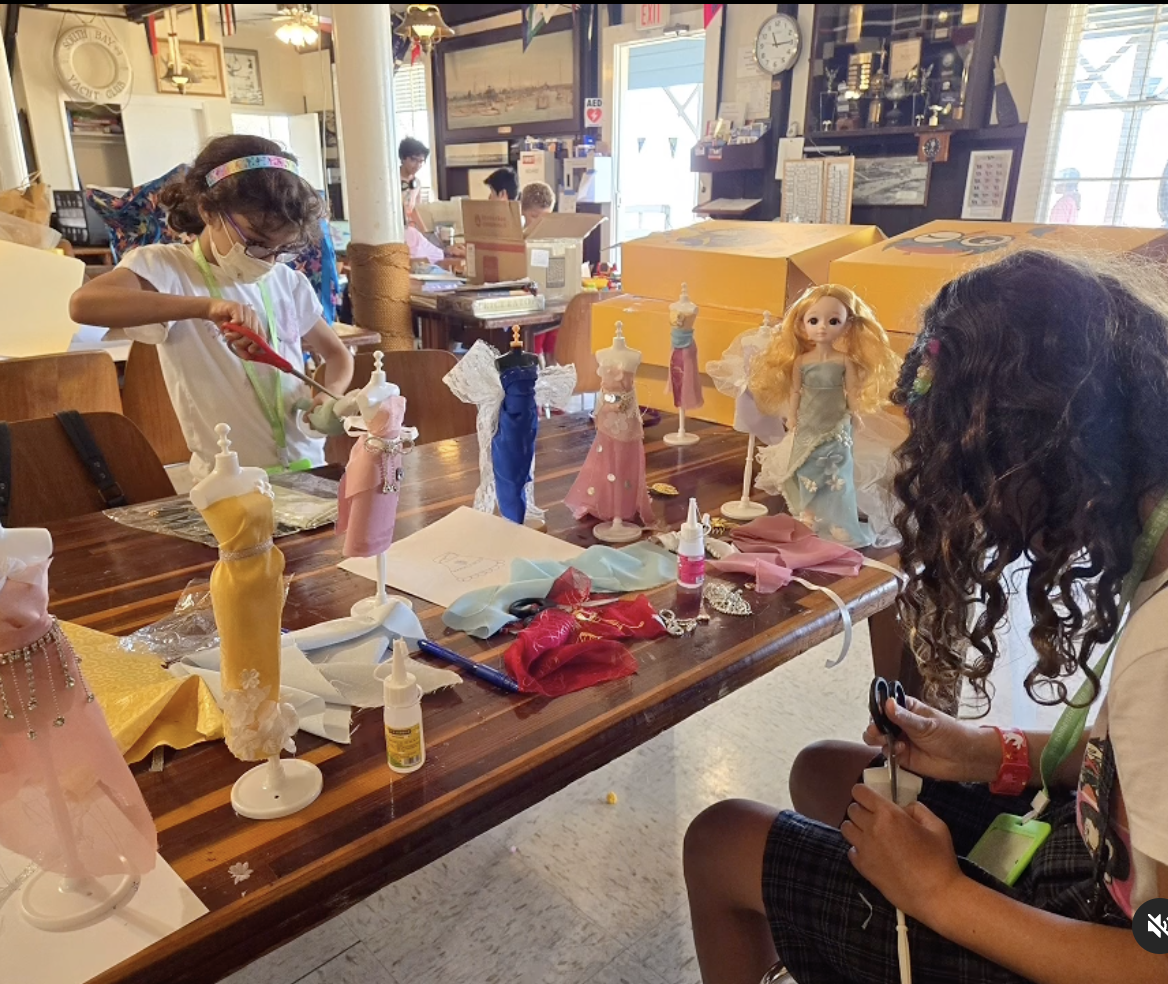 Children crafting doll clothes at a table with dress mannequins and art supplies in a classroom.