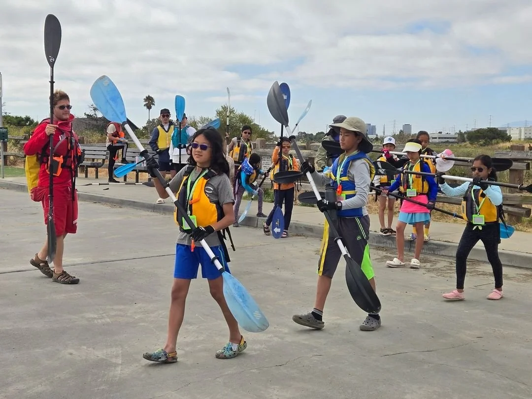 Group of children and adults wearing life jackets, carrying kayak paddles, walking on a paved path, and preparing for a kayaking activity.