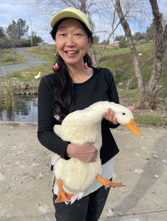 Woman smiling while holding a white duck outdoors near a pond and trees.