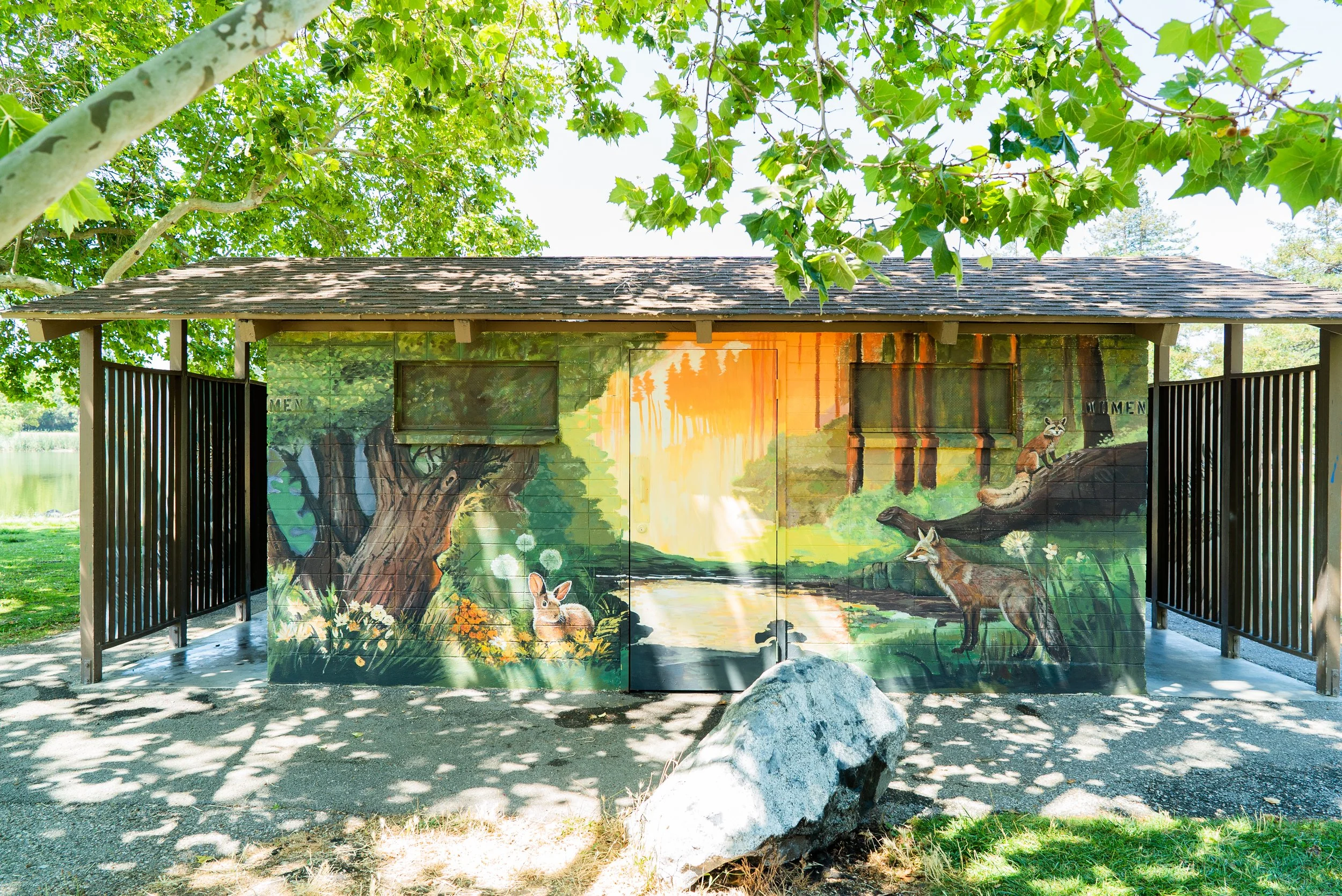Park restroom building with a nature-themed mural depicting a forest scene with animals, including foxes, a rabbit, and a sunset, surrounded by green trees and foliage.