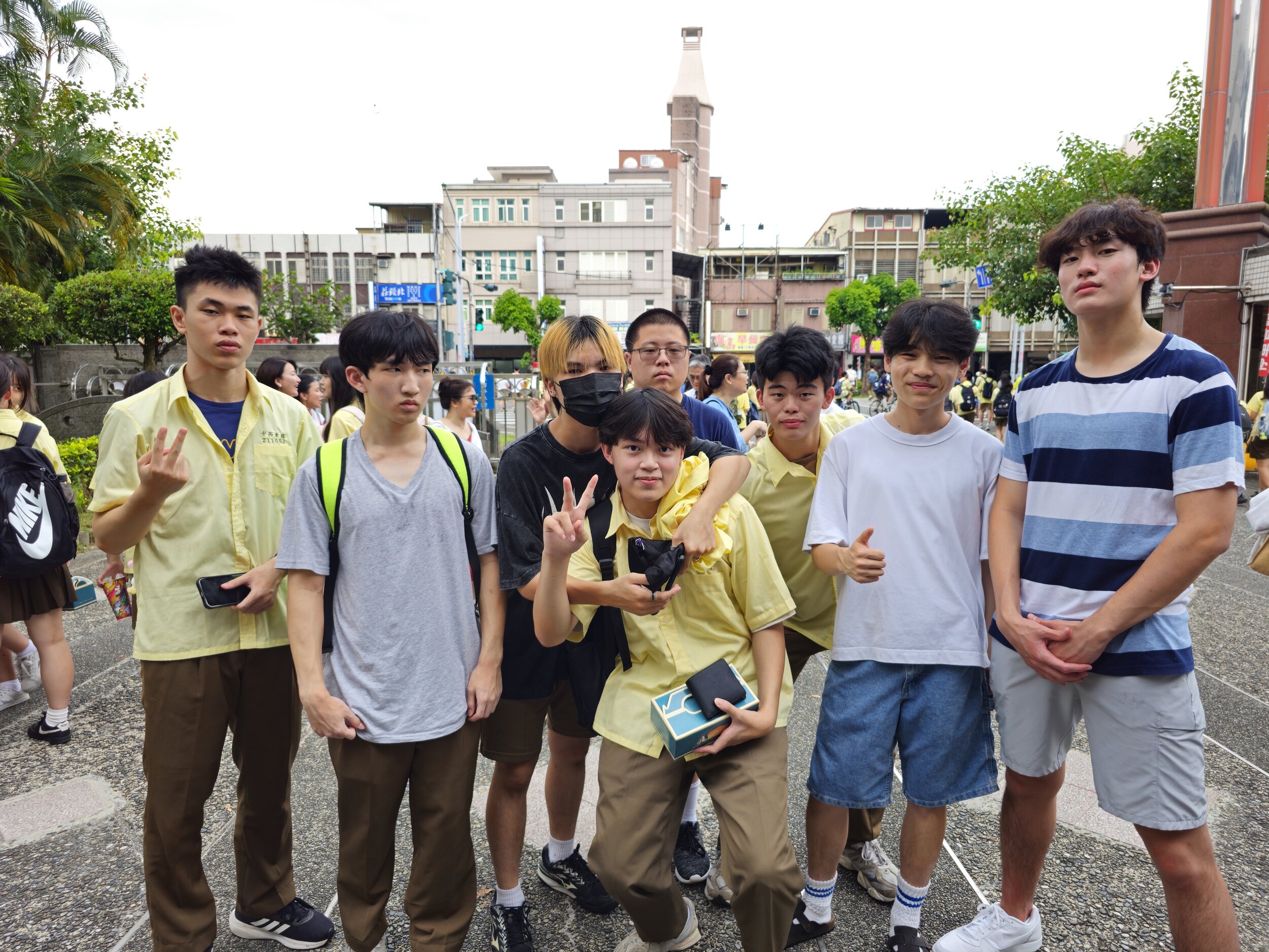 Group of young people posing for a photo outdoors, some wearing yellow shirts, with a school building in the background. Two people are making peace signs, and one is giving a thumbs-up. It appears to be a casual or school-related gathering.