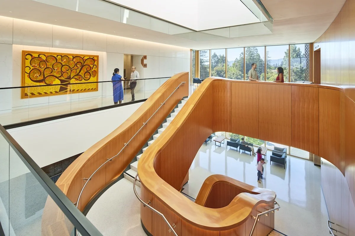 Interior of a modern building with a wooden staircase and a glass railing. There are people talking and sitting near large windows showing trees outside, with a colorful abstract art on the wall.