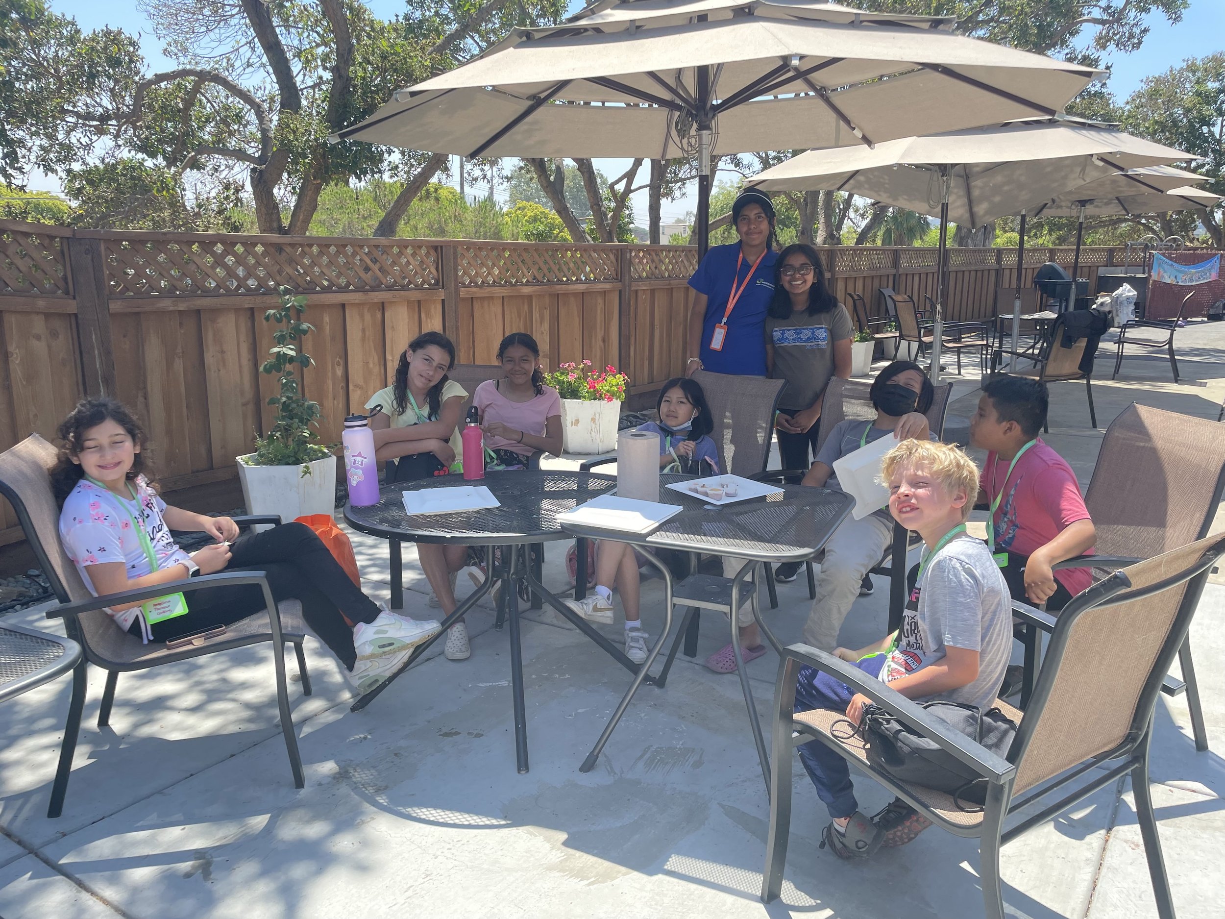 A group of children sitting and standing around a patio table under umbrellas. The setting appears to be outdoors with a wooden fence and trees in the background. Some of the children are seated in chairs, while others are standing, and they all seem