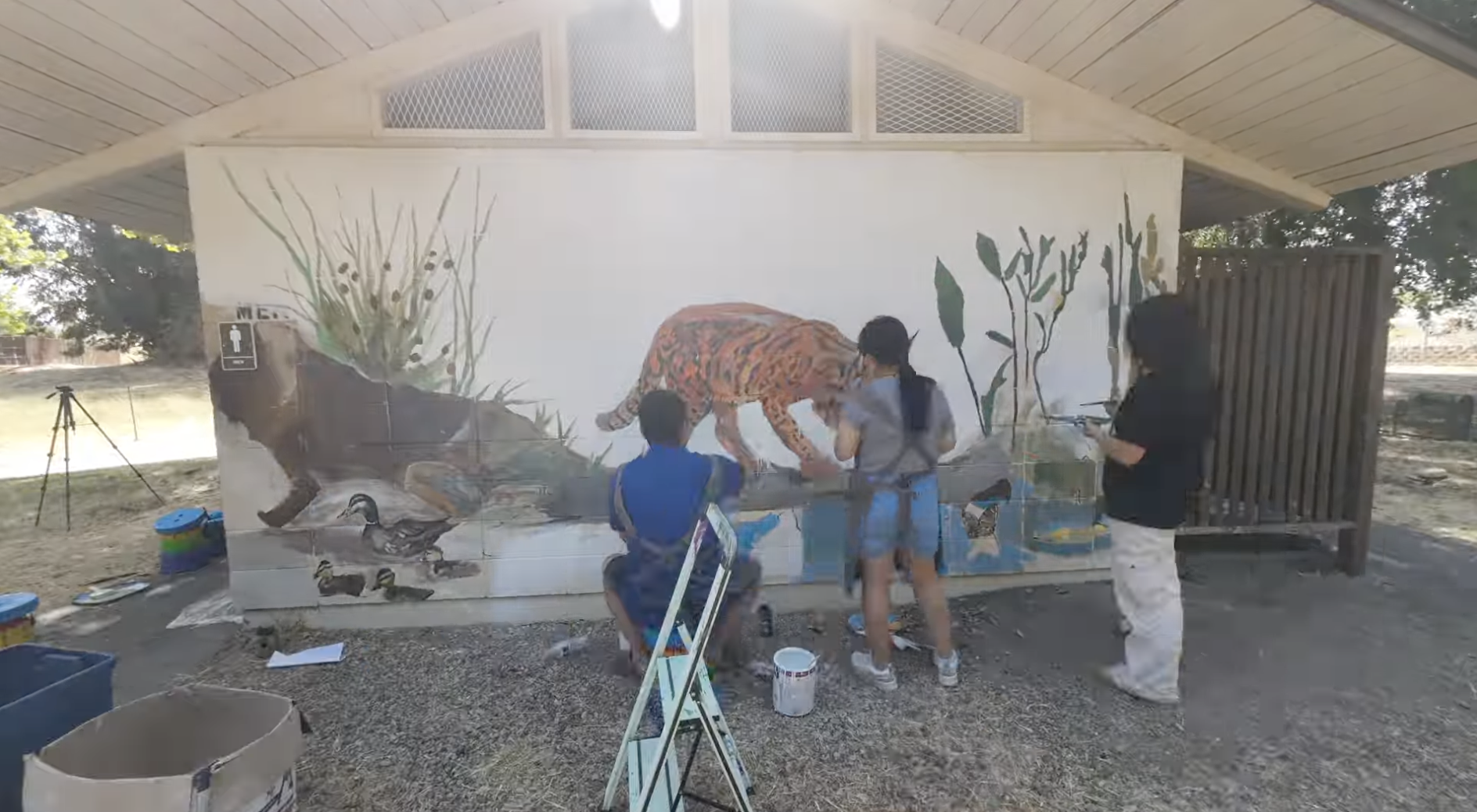 Three people painting a mural on a building wall, depicting a tiger, ducks, and vegetation.