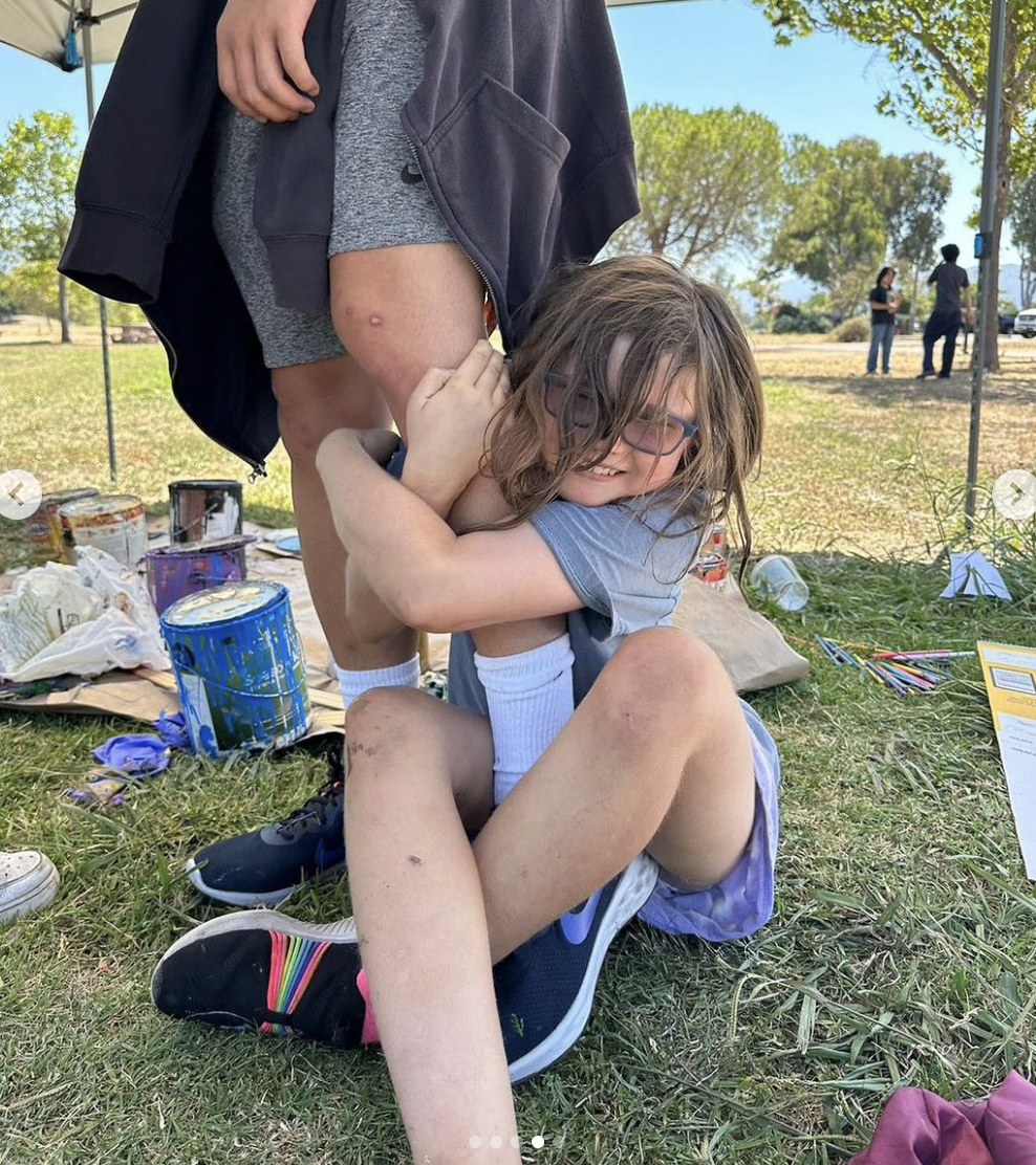 Child sitting on grass hugging a person's leg, surrounded by paint cans and art supplies, outdoors on a sunny day.