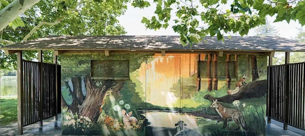 A painted mural depicting wildlife, including deer and rabbits, on a wooden structure near a lake, surrounded by trees.