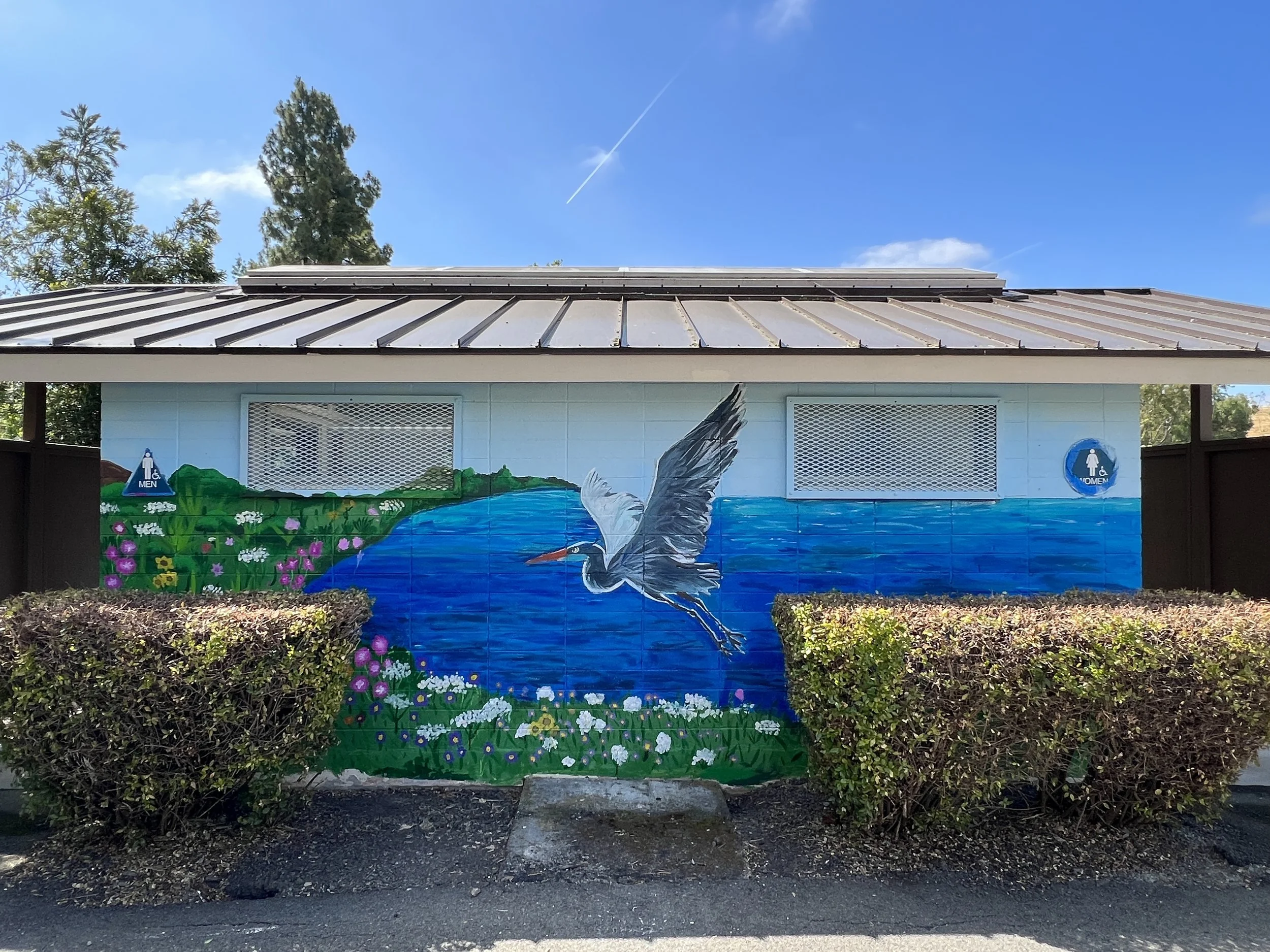 Mural on building wall depicting a heron flying over a lake with surrounding greenery and flowers, located between men's and women's restroom signs.