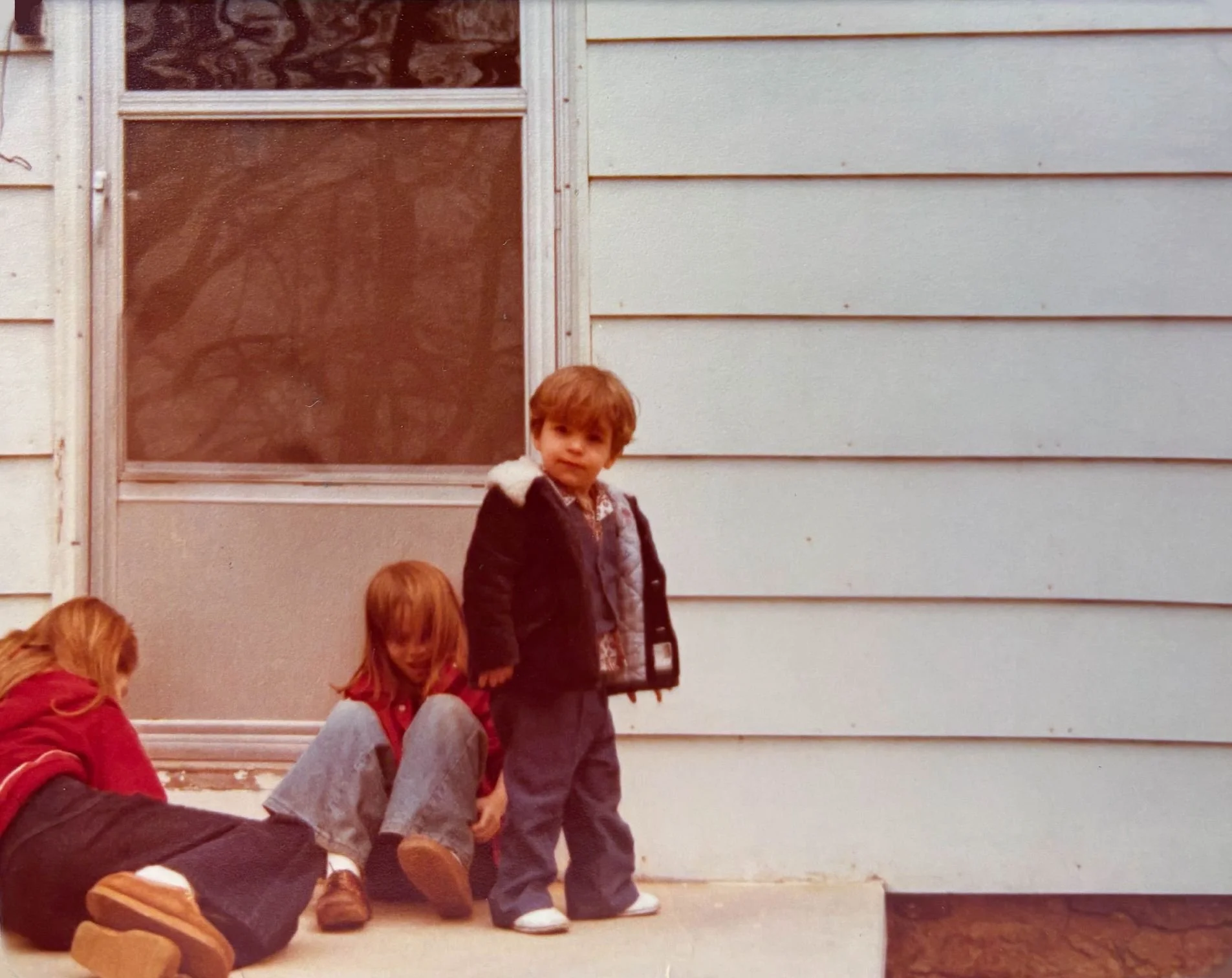 a young boy and his sisters on the front stoop of a modest home, taken with an old film camera. Theres a soft amber glow of age over the photograph