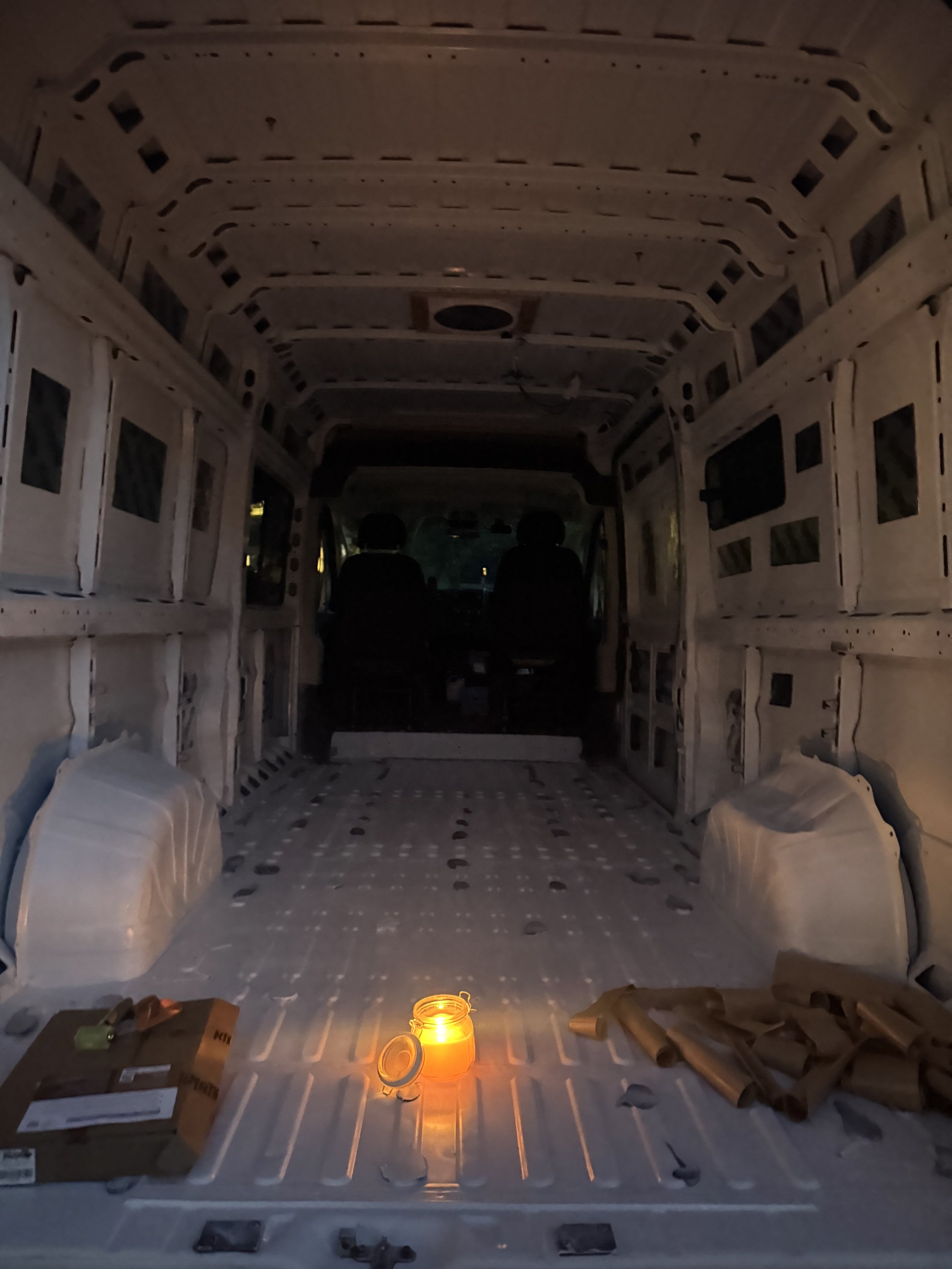 Interior of a stripped out van in the dark with a dingle lit candle glowing orange on the floorboard.cardboard boxes litter the edges of the frame