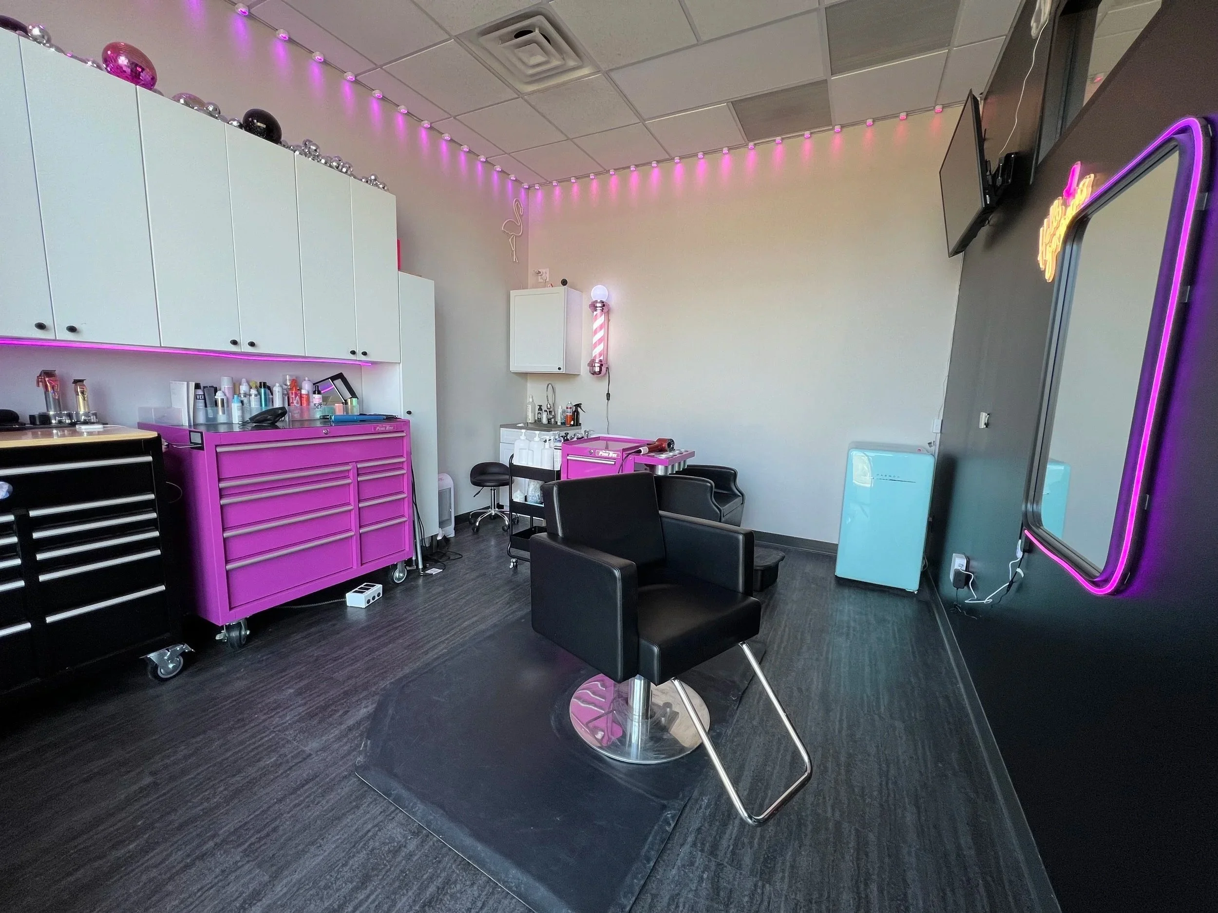 Whole room photo of Embee Hair Salon suite in Davenport, Iowa. Fun atmosphere featuring neon lights, a tv, comfortable shampoo stations and a hot pink tool box to hold all the tools for great haircuts. Fully stocked color bar.