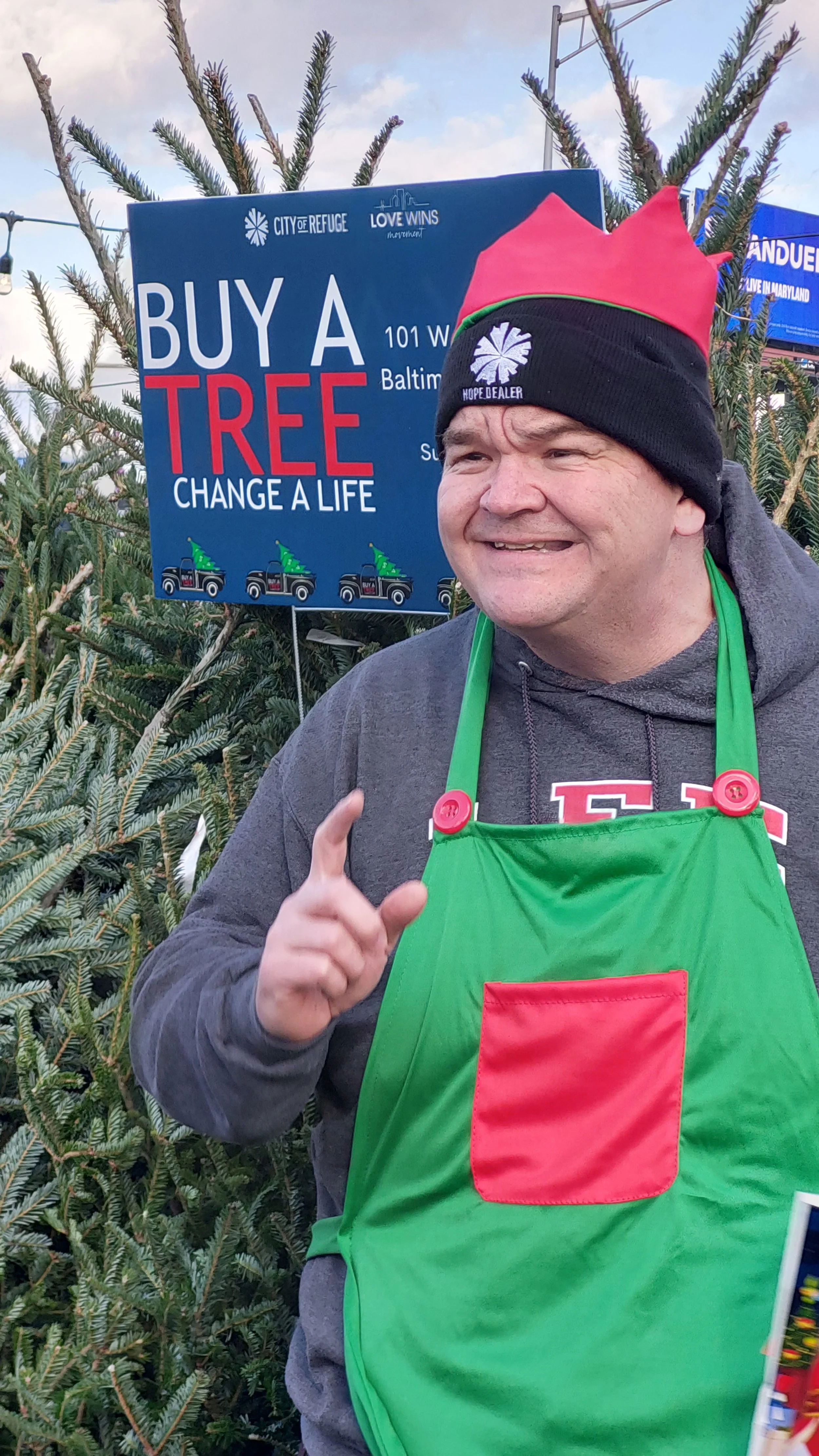 Our Executive Director wearing a green apron and a red and black Christmas hat standing in front of a Christmas tree and a sign that says 'Buy a Tree, Change a Life'.