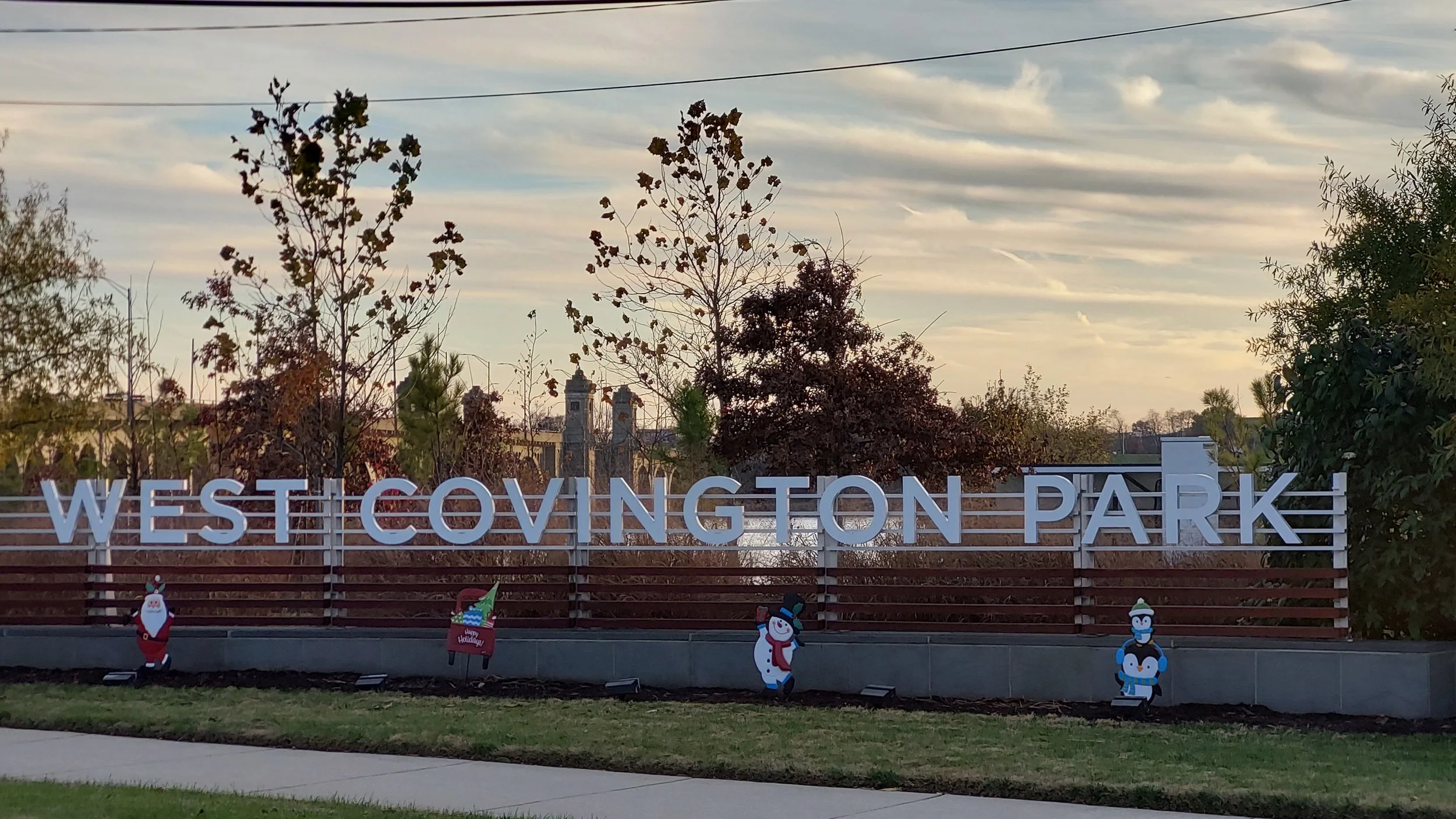 Sign reading 'West Covington Park' with holiday decorations including Santa, snowmen, and a penguin in front of trees and a partly cloudy sky.