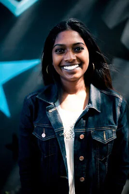 A young woman with dark hair and dark eyes smiling, wearing a denim jacket and a white top, standing indoors with an abstract blue background.