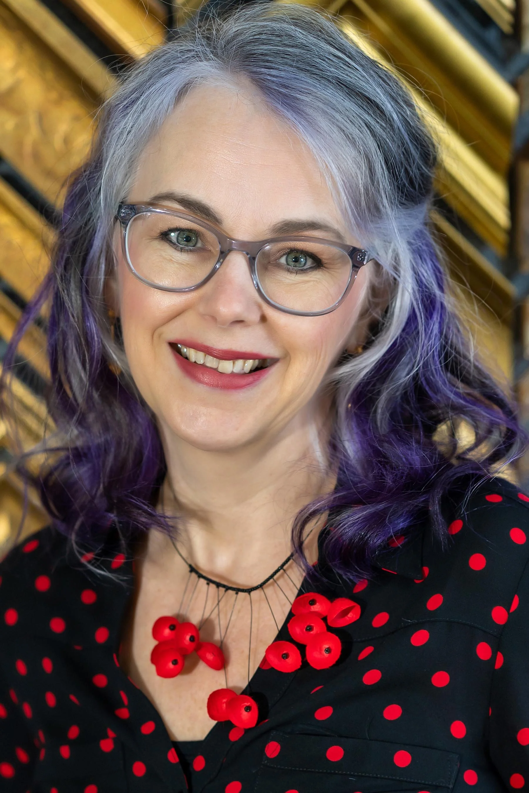A woman with glasses, silver and purple hair, smiling, wearing a black dress with red polka dots and a red necklace, against a wooden background.