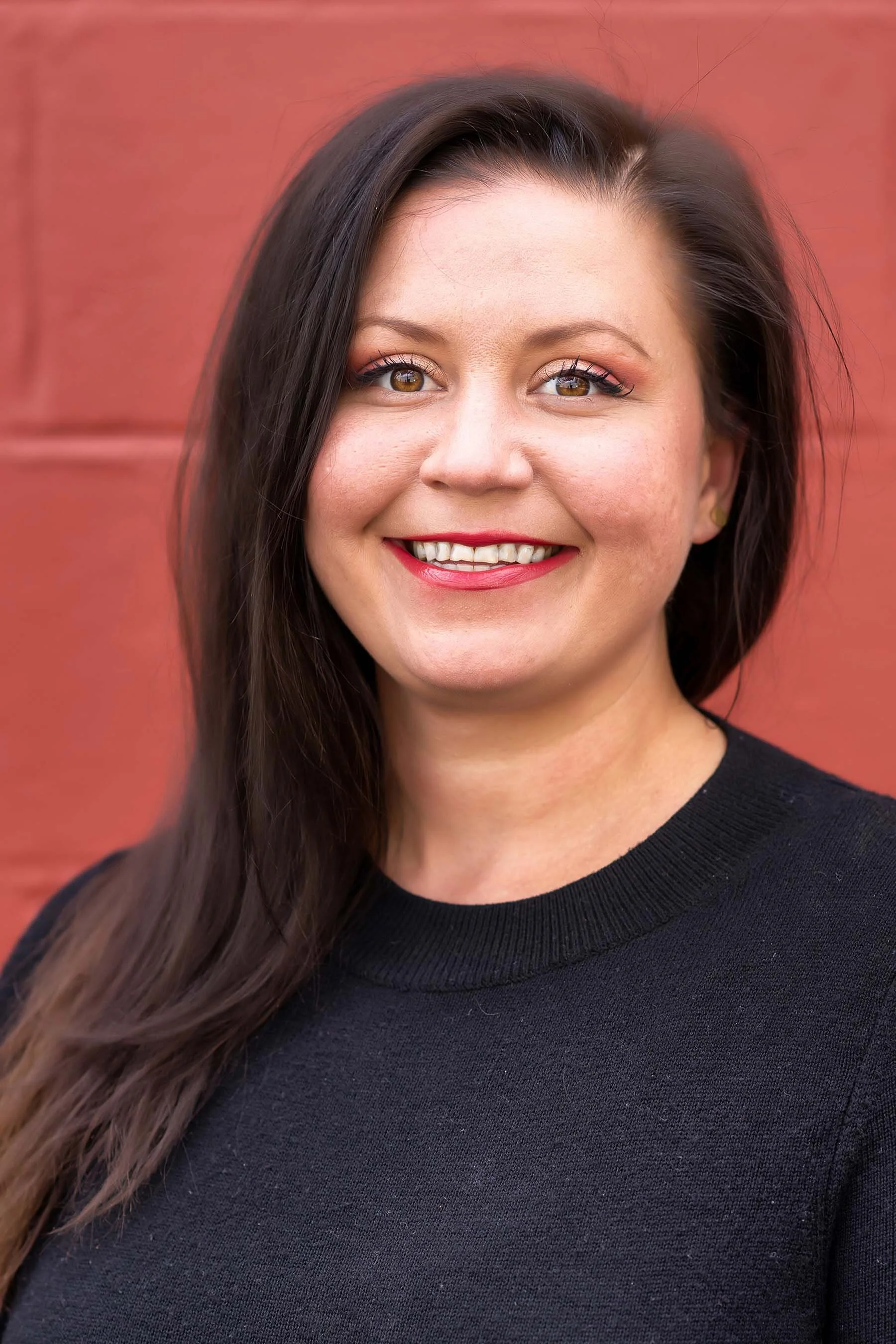 A woman with long dark brown hair, wearing a black top, smiling in front of a reddish-orange background.