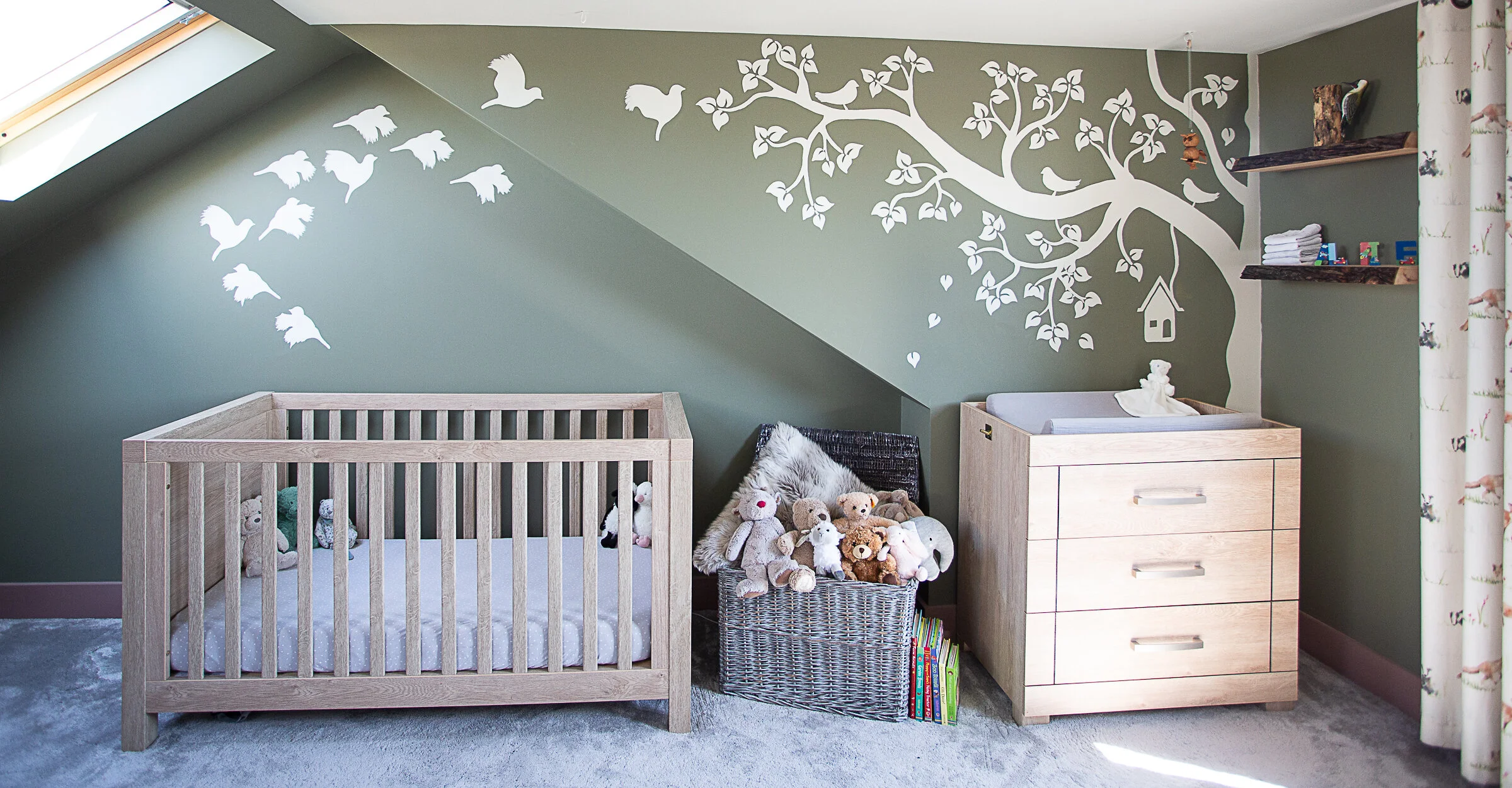 A Modern country design. Featuring a large autumnal tree and flying birds set with a birdcage. The mural is green, with designs in a pink and off-white colour combination. In front of the mural is a crib, a toybox with bears, and a drawer.