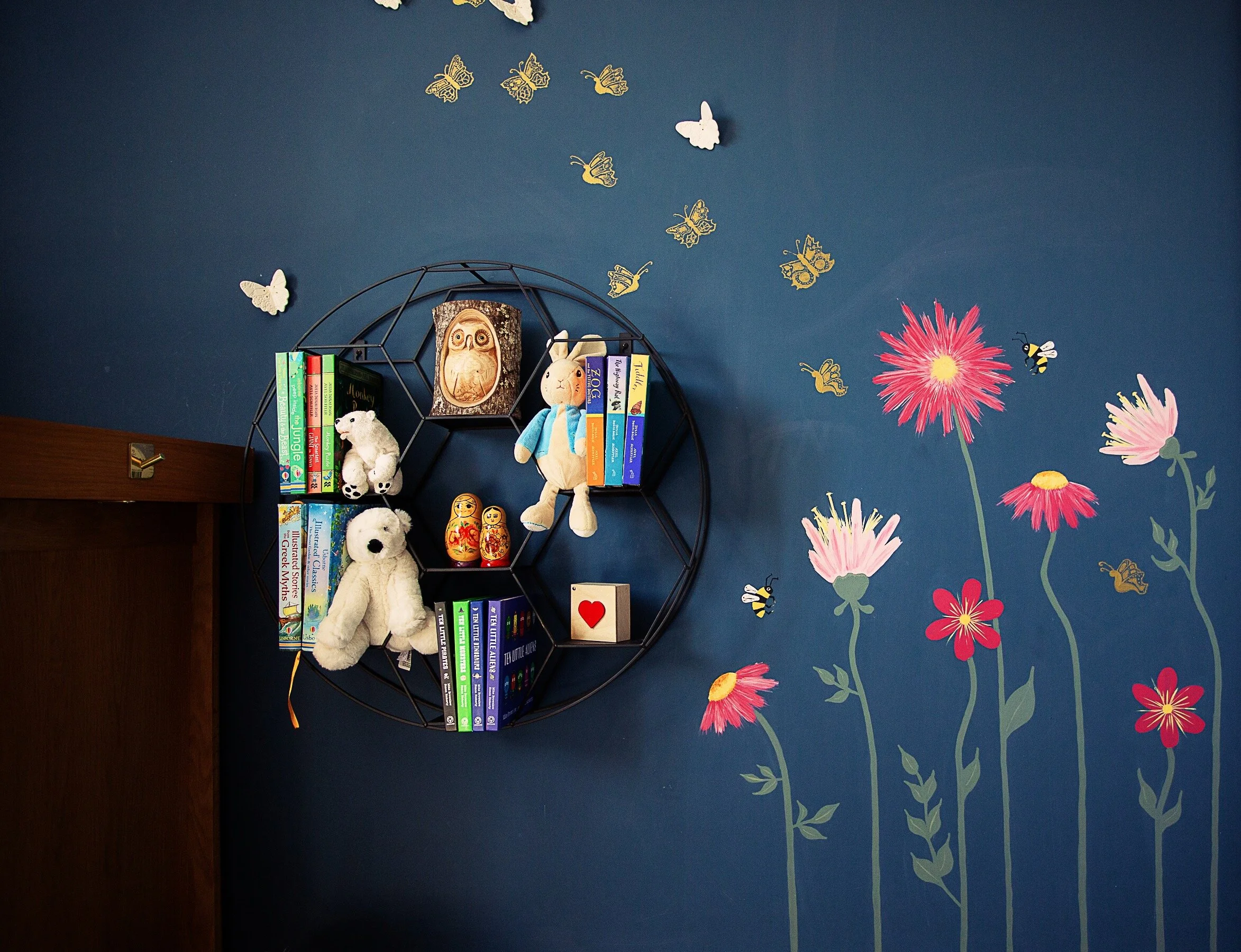 A closeup of the fuchsia pink wildflowers on the dark blue wall. To the left of the flowers is a circular shelf with stuffed animals and small books.