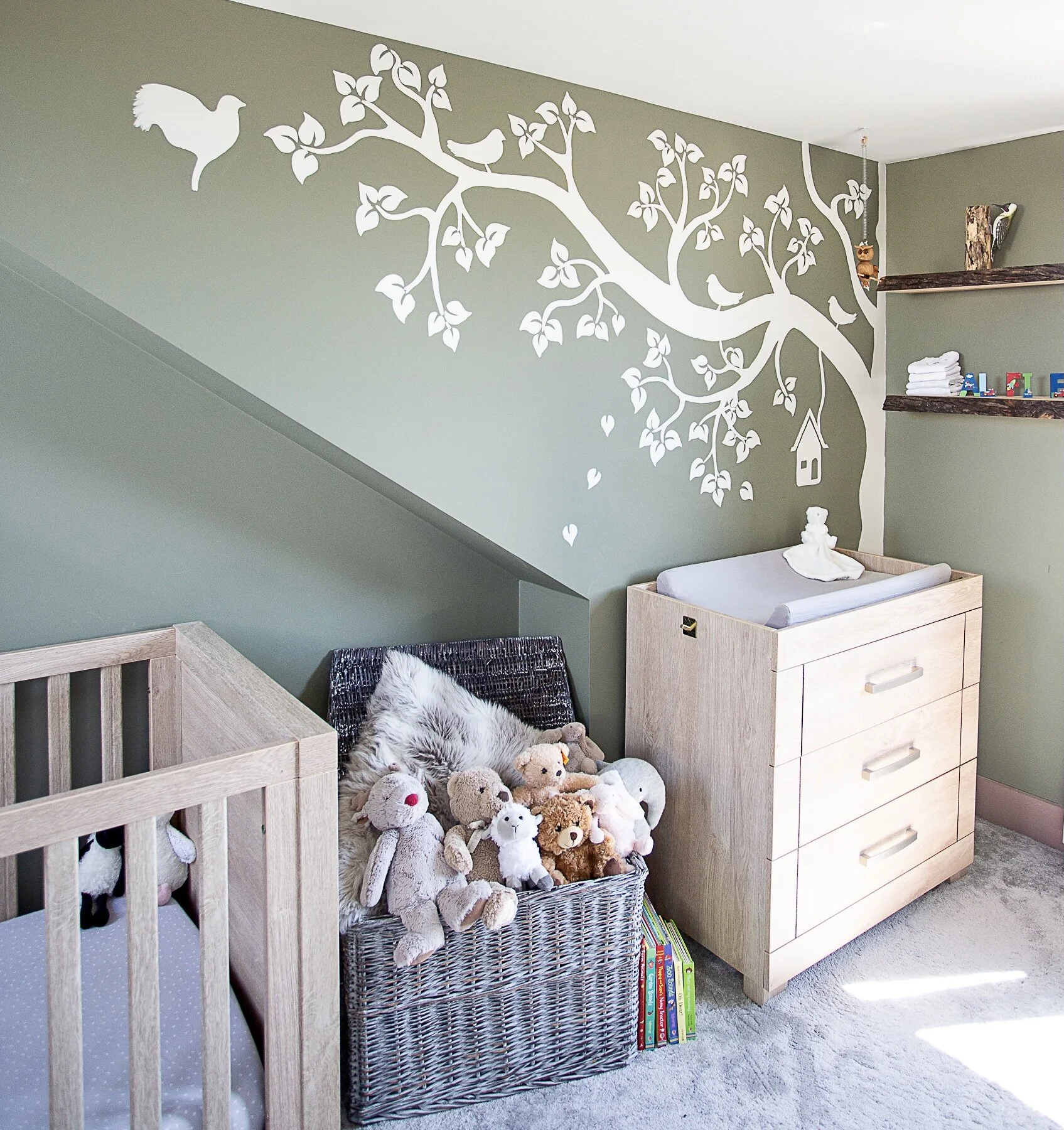A depiction of the far right corner of the nursery. The mural has a modern country design featuring a large autumnal tree and flying birds set with a birdcage. The mural is green, with designs in a pink and off-white colour combination. In front of t