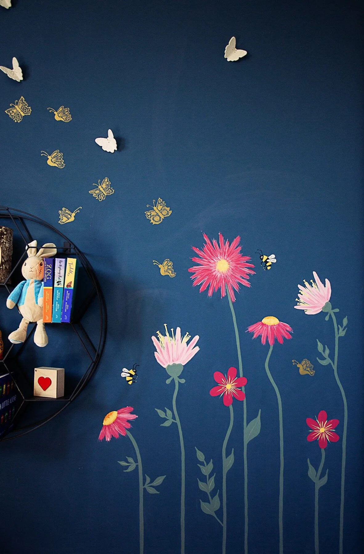 A closeup of the fuchsia pink wildflowers on the dark blue wall. To the left of the flowers is a circular shelf with stuffed animals and small books.