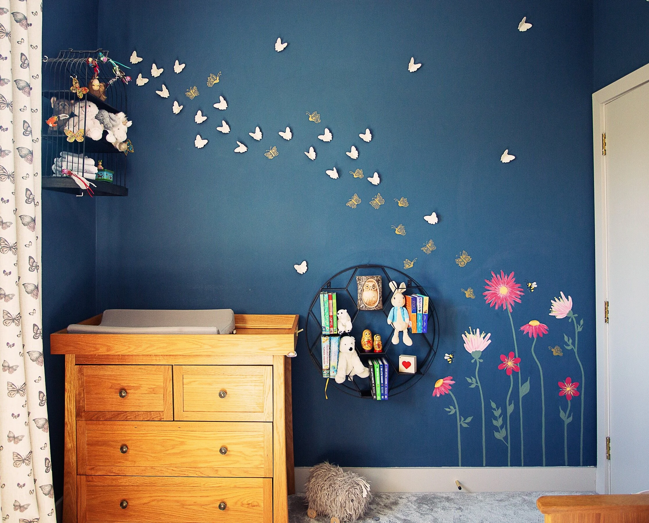 White porcelain butterflies depart from fuchsia pink wildflowers on a dark blue wall. In front of the mural are two shelves and  a drawer. 