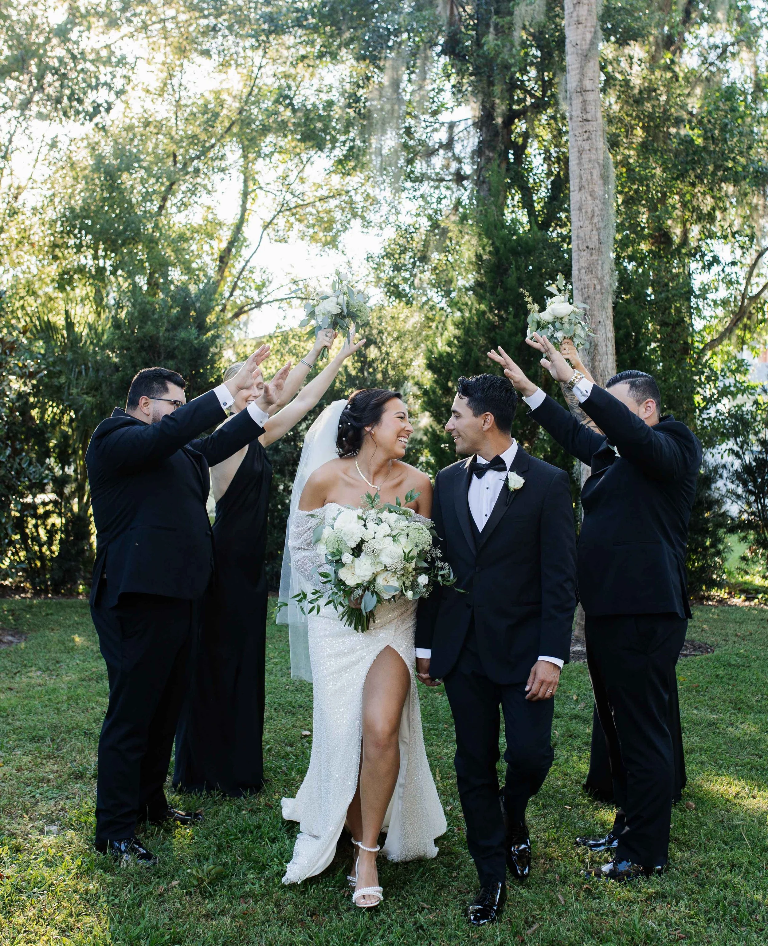 Wedding couple walking hand-in-hand beneath arched hands of five people holding bouquets, in an outdoor garden setting with trees and sunlight.