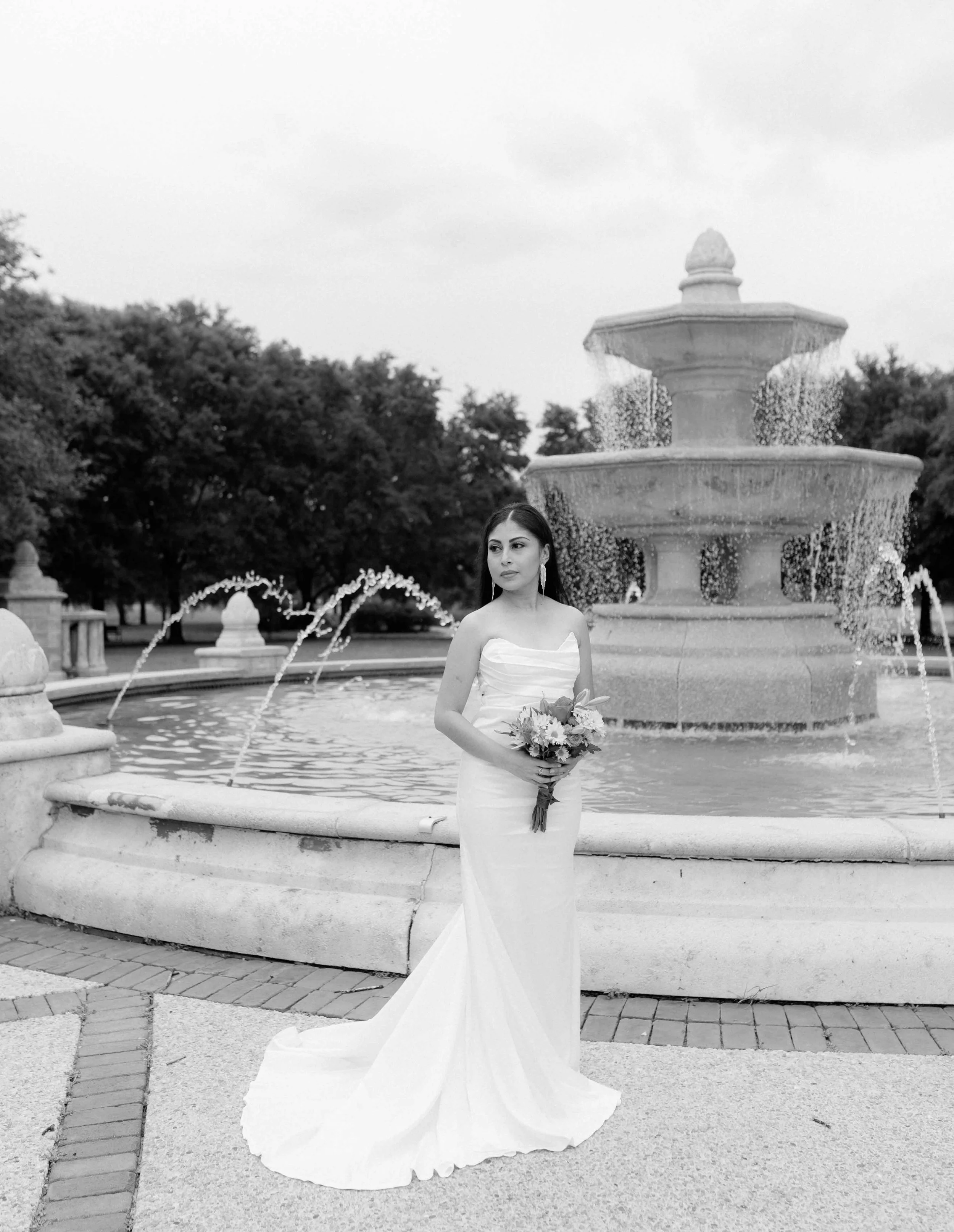 A bride in a strapless wedding gown holding a bouquet stands in front of a multi-tiered fountain in a park.