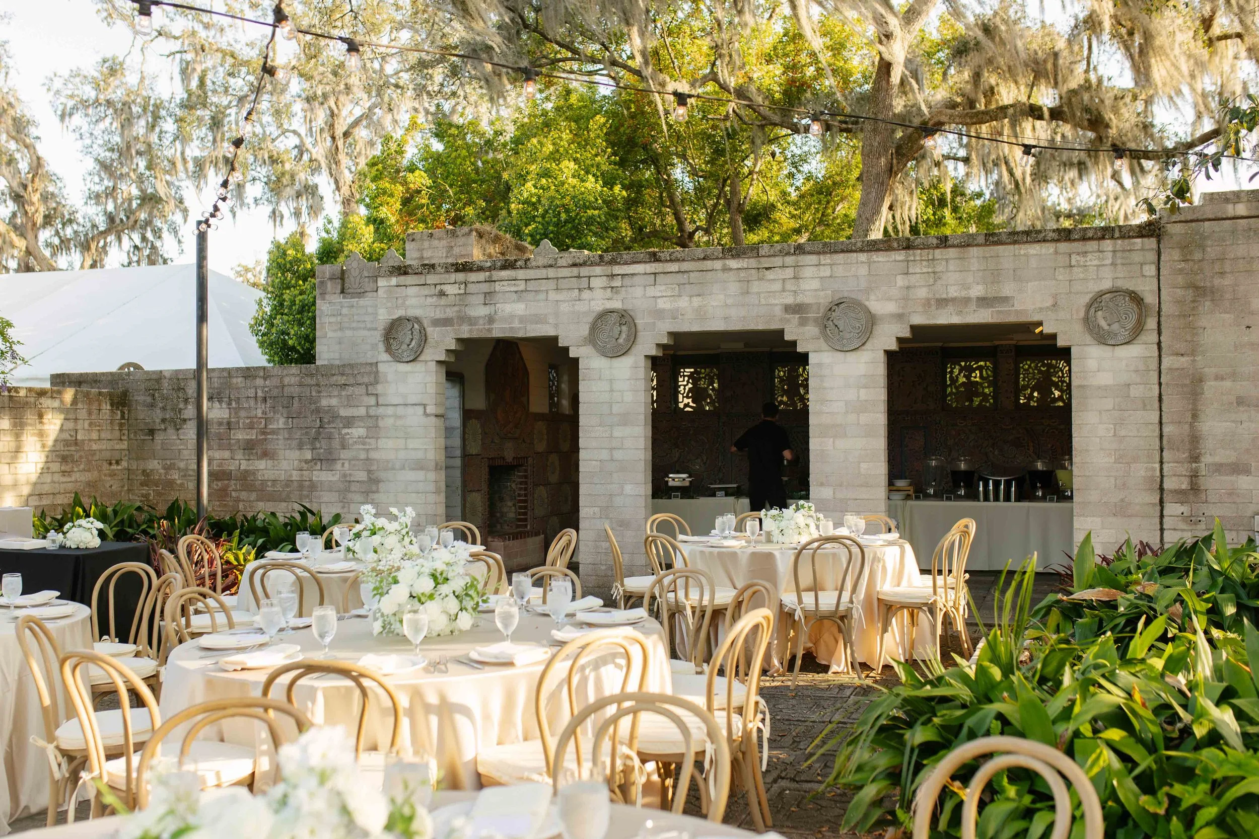 Outdoor event setup with round tables, white tablecloths, floral centerpieces, and chairs, in front of a stone structure with decorative plaques, surrounded by greenery and string lights.