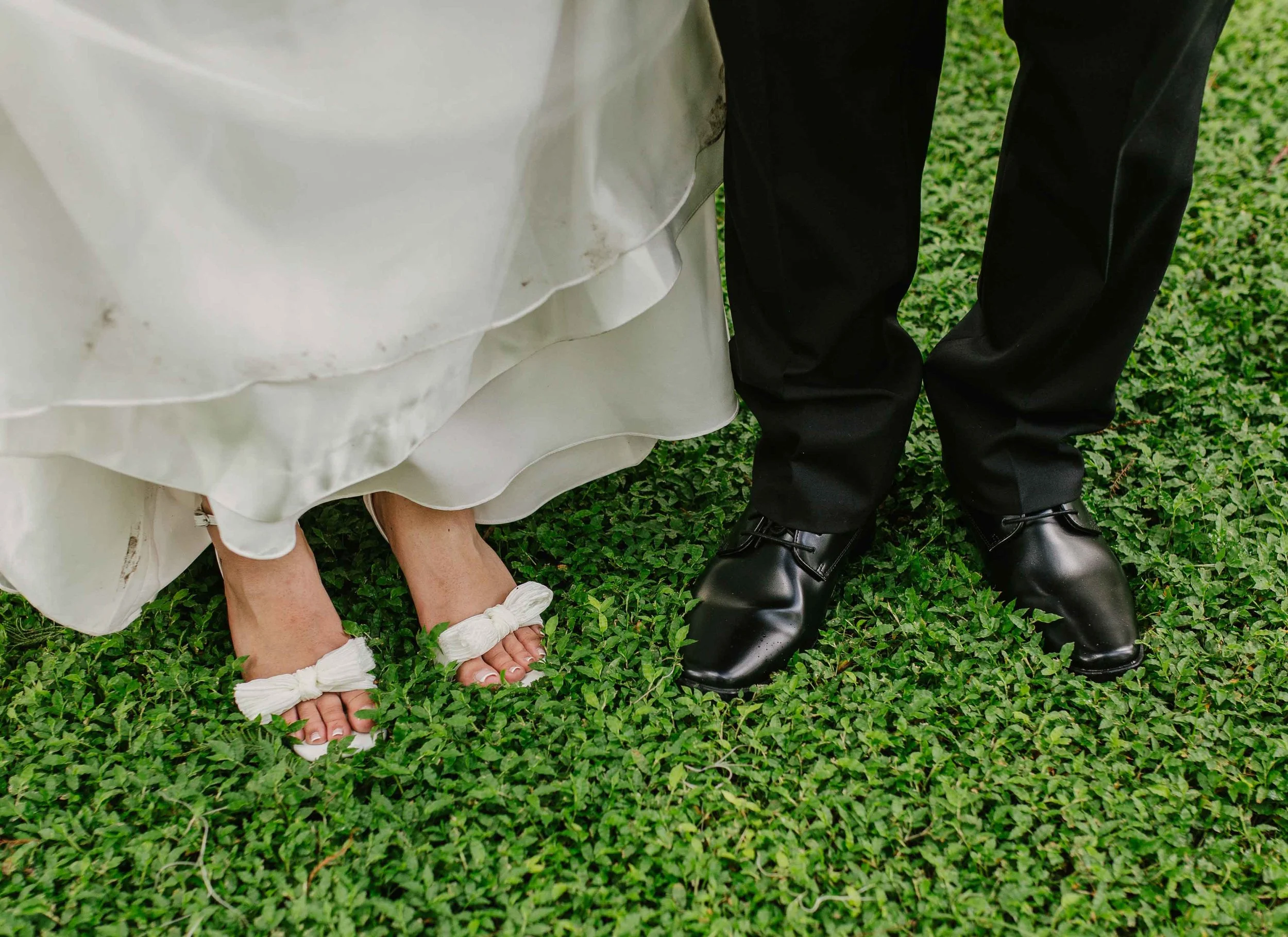 Close-up of wedding couple's feet on green grass; woman wearing white sandals with bows and man in black dress shoes.