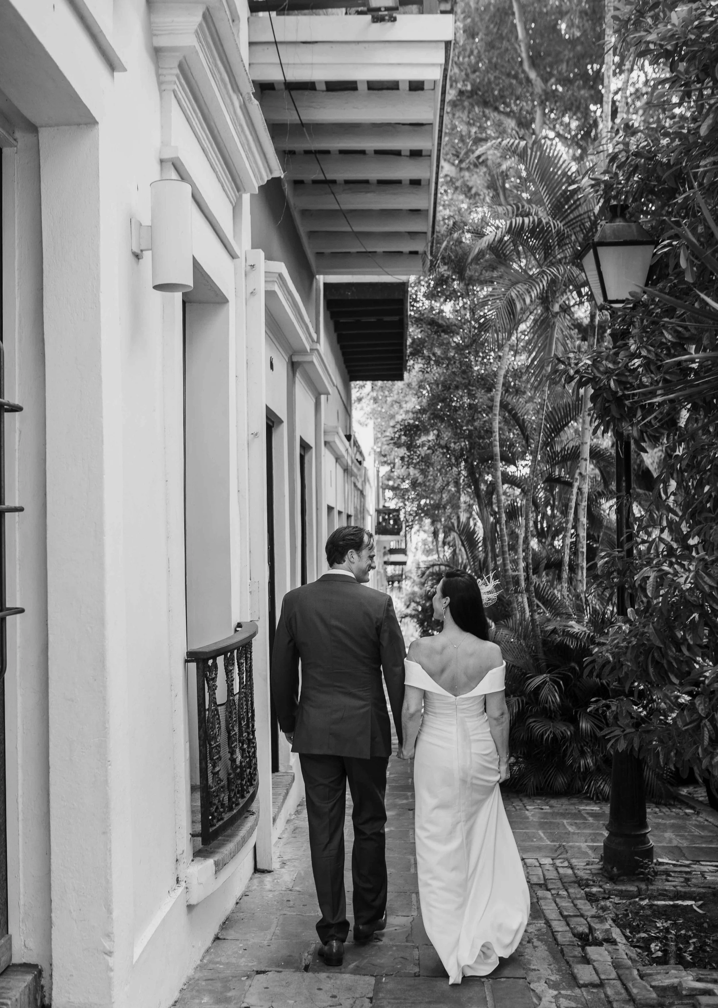 A black and white photo of a couple in wedding attire walking down a narrow alleyway with lush greenery on the side.