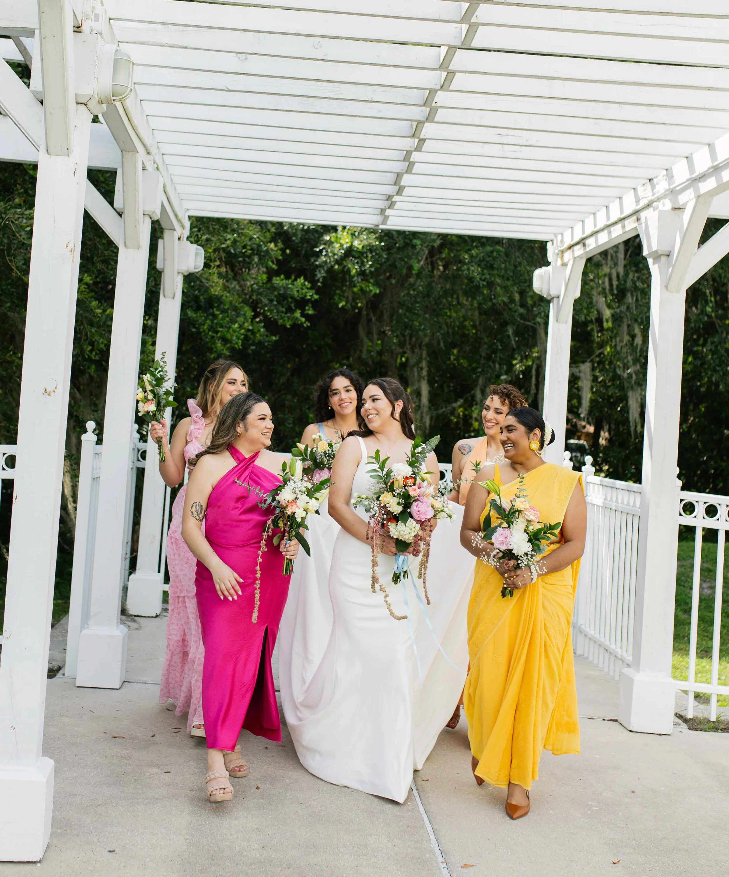 Group of diverse women in colorful dresses holding bouquets, walking outdoors under a white pergola with green trees in the background, celebrating a wedding.