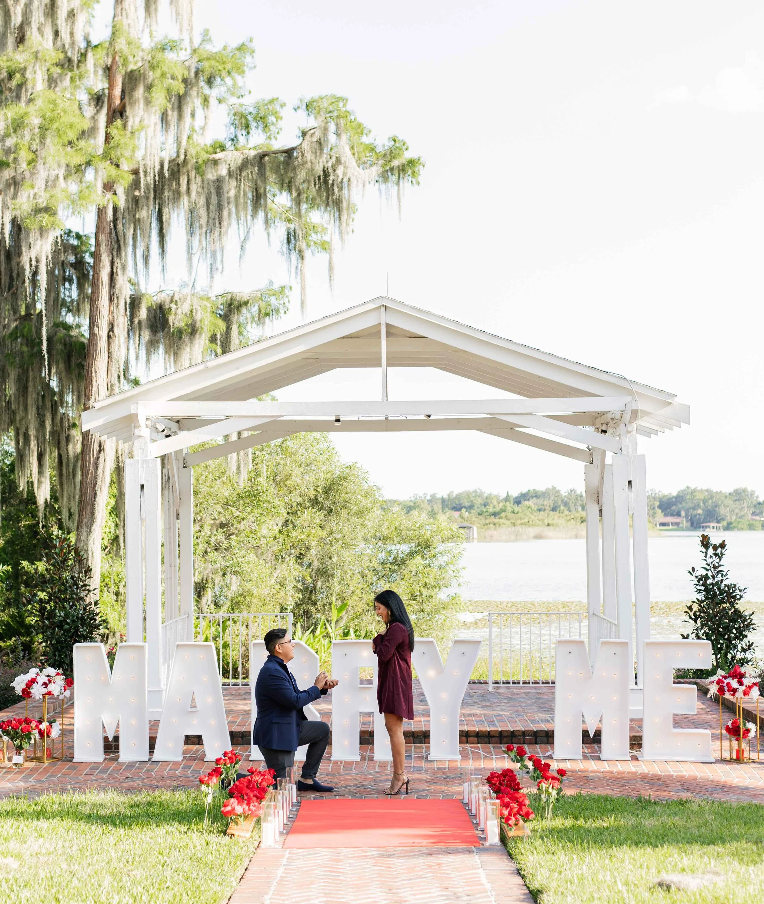 A couple proposes during a romantic outdoor setup with large illuminated letters spelling 'MARRY ME' near a lakeside, decorated with red roses and a red carpet walkway.