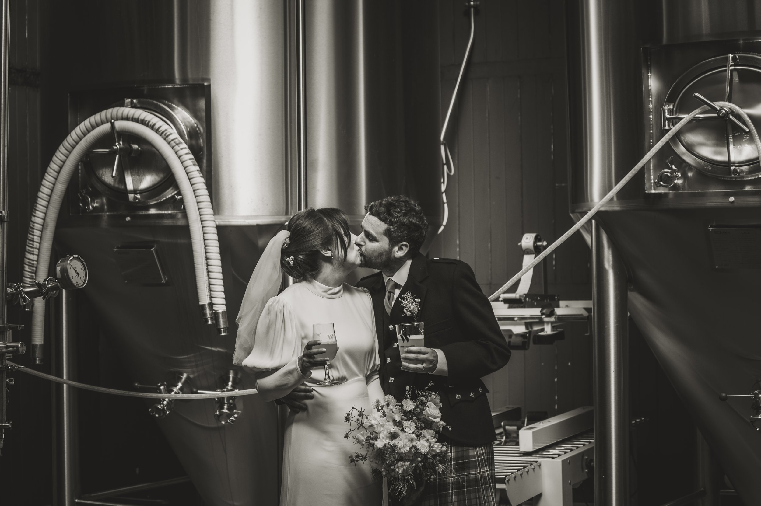 Couple in wedding attire kissing in a brewery among large tanks, holding beer glasses.
