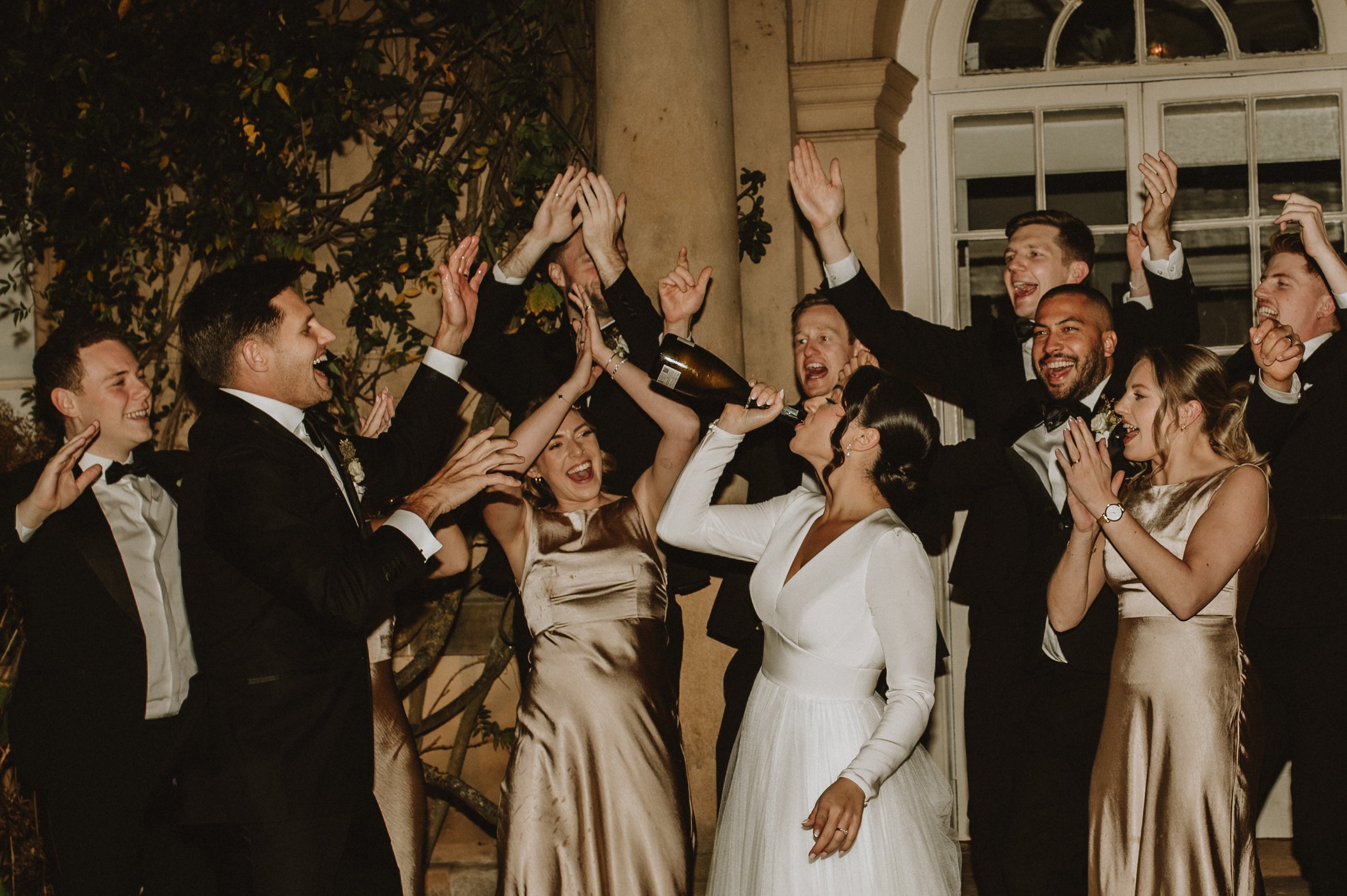 Group celebrates a wedding, with a bride drinking from a champagne bottle, surrounded by joyful guests in formal attire at night, raising hands in celebration.
