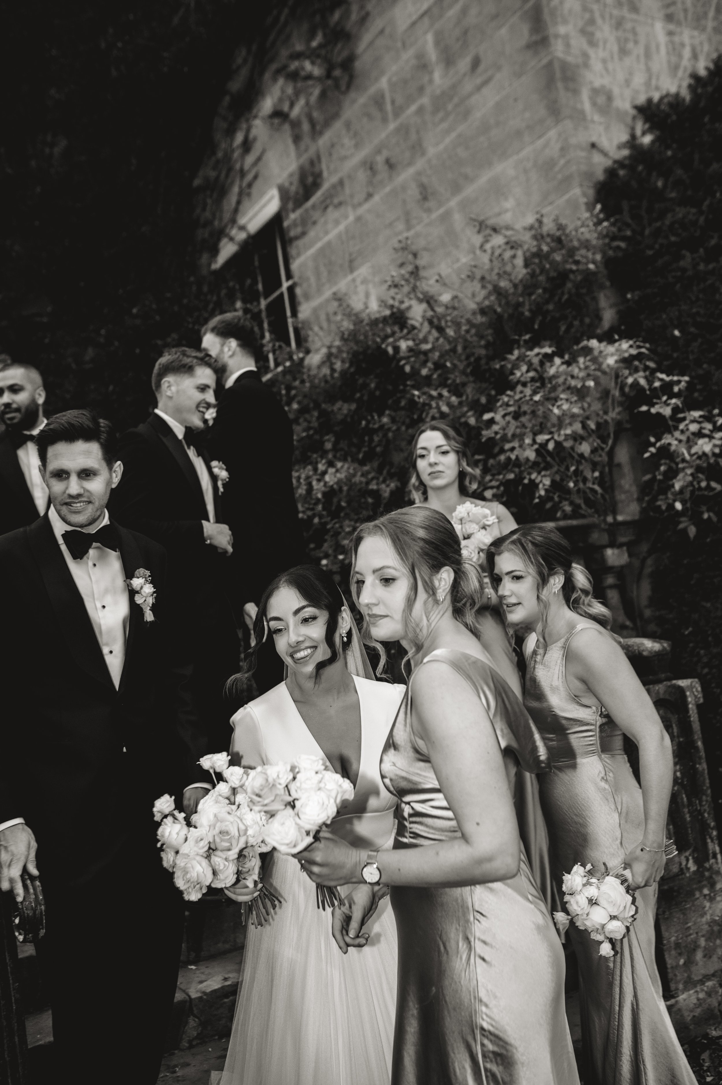 Black and white photo of a wedding party, including a bride in a gown with flowers, bridesmaids in dresses, and groomsmen in tuxedos, standing on steps outside.